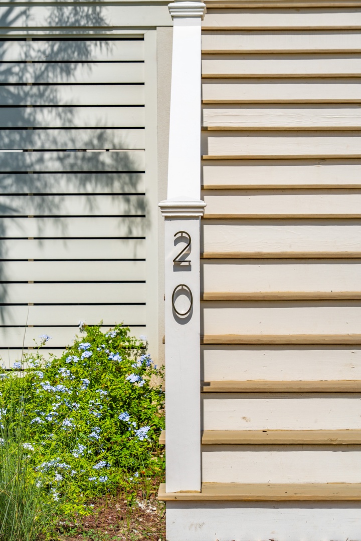 The entrance to this rental property features a welcoming facade with house number 20 clearly displayed on a white pillar, surrounded by charming blue flowers and natural plantings.