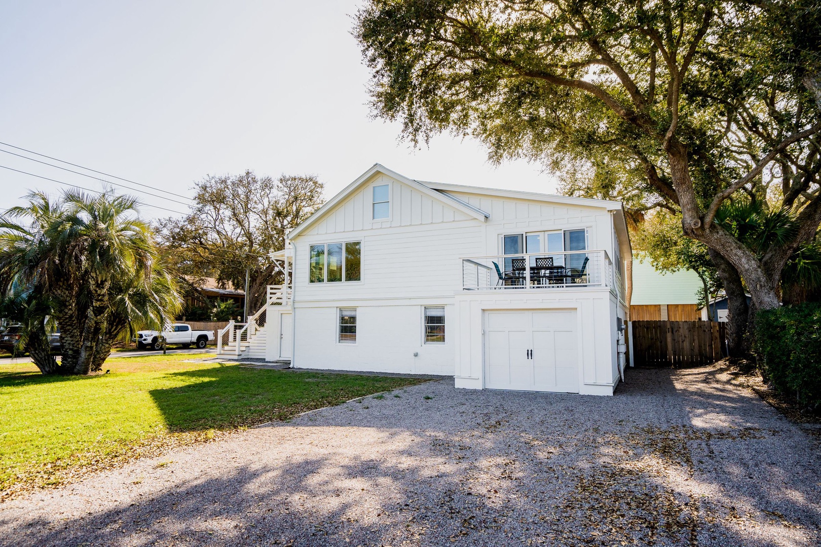 Charming white coastal home with garage parking and upper deck, nestled among tropical palms in a peaceful neighborhood setting.