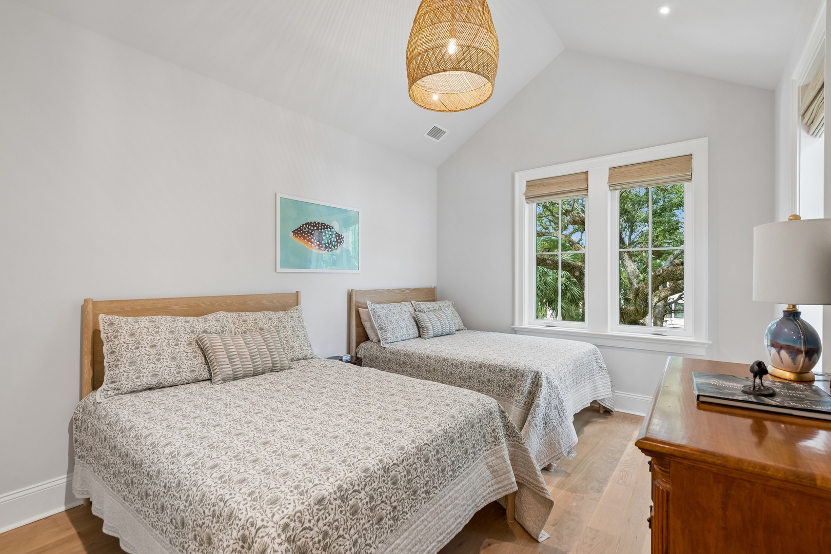 Your bright guest bedroom offers two plush beds beneath vaulted ceilings and a charming woven pendant light, while natural light pours in through French doors framing lush garden views.