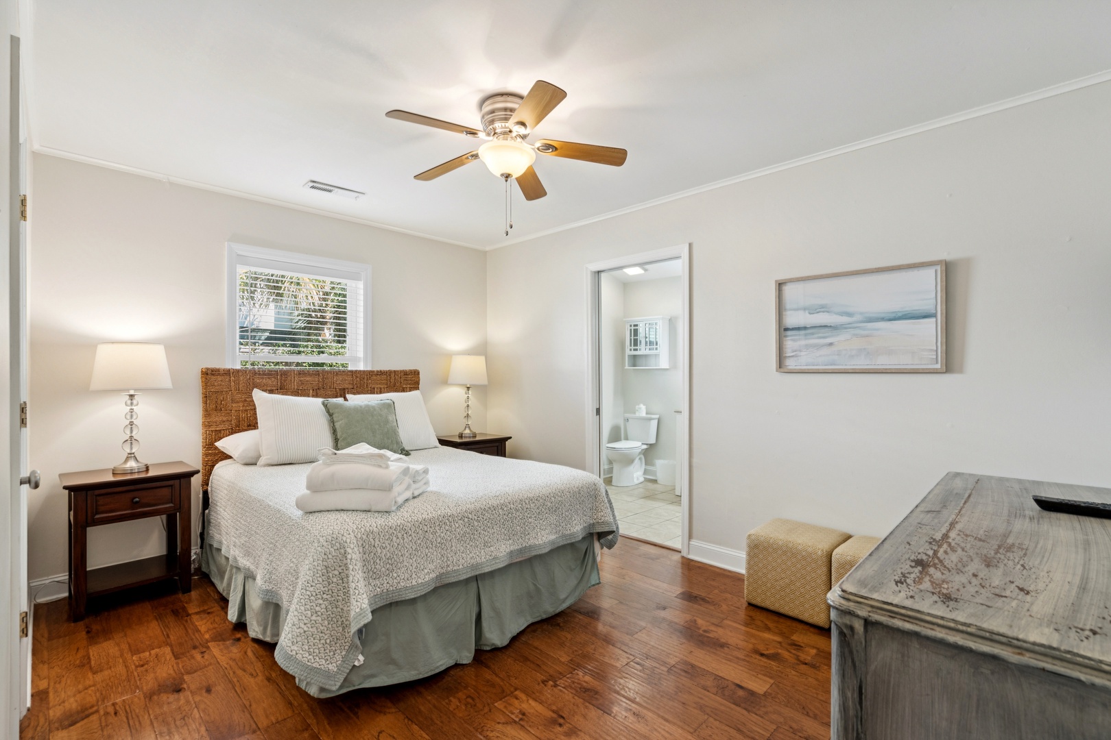 Unwind in your peaceful bedroom featuring coastal artwork, soft linens, and gleaming hardwood floors beneath a gentle ceiling fan.