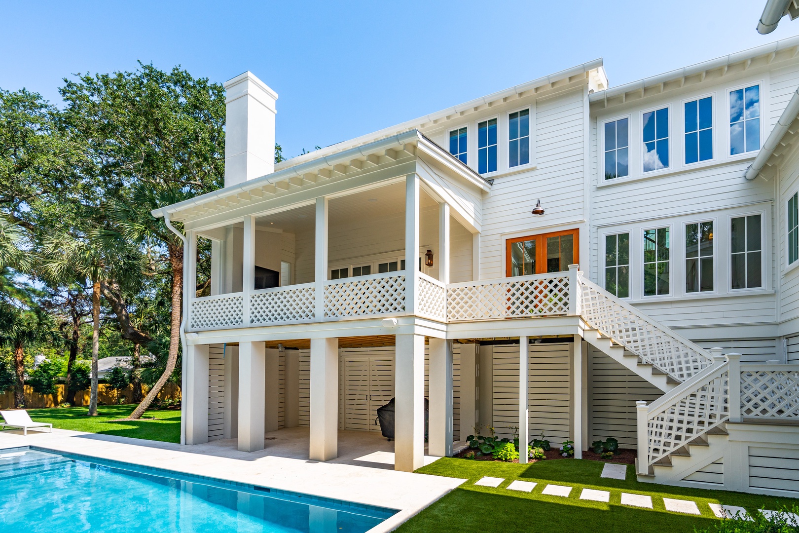 A stunning white coastal property showcases a covered porch with lattice railings, an expansive pool, and lush landscaping framed by swaying palms and a bright blue sky.