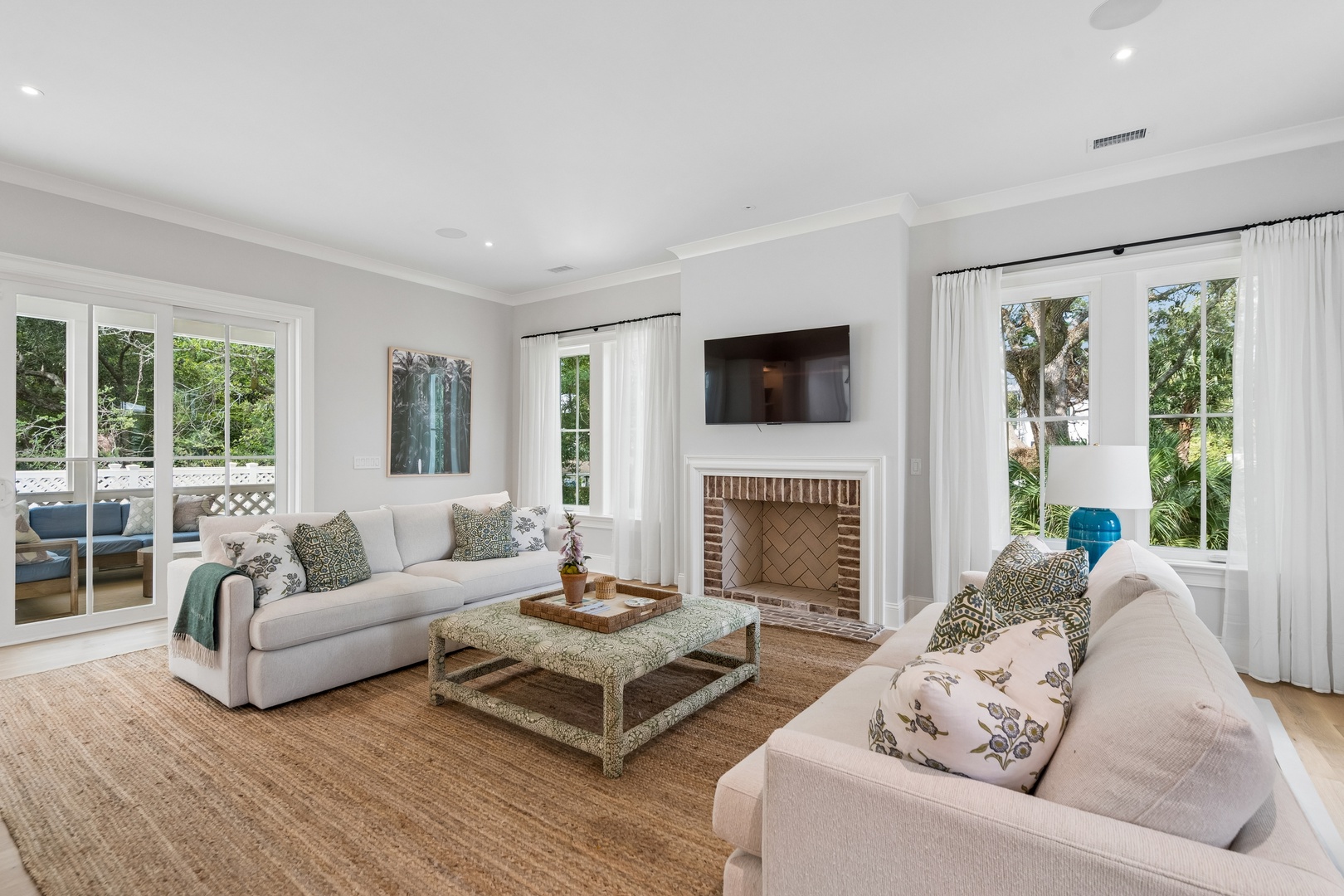 Settle into your bright, welcoming living room where soft sofas invite relaxation and a decorative fireplace anchors the space. Large windows and patio doors frame lush greenery outside, filling the room with natural light and peaceful garden views.