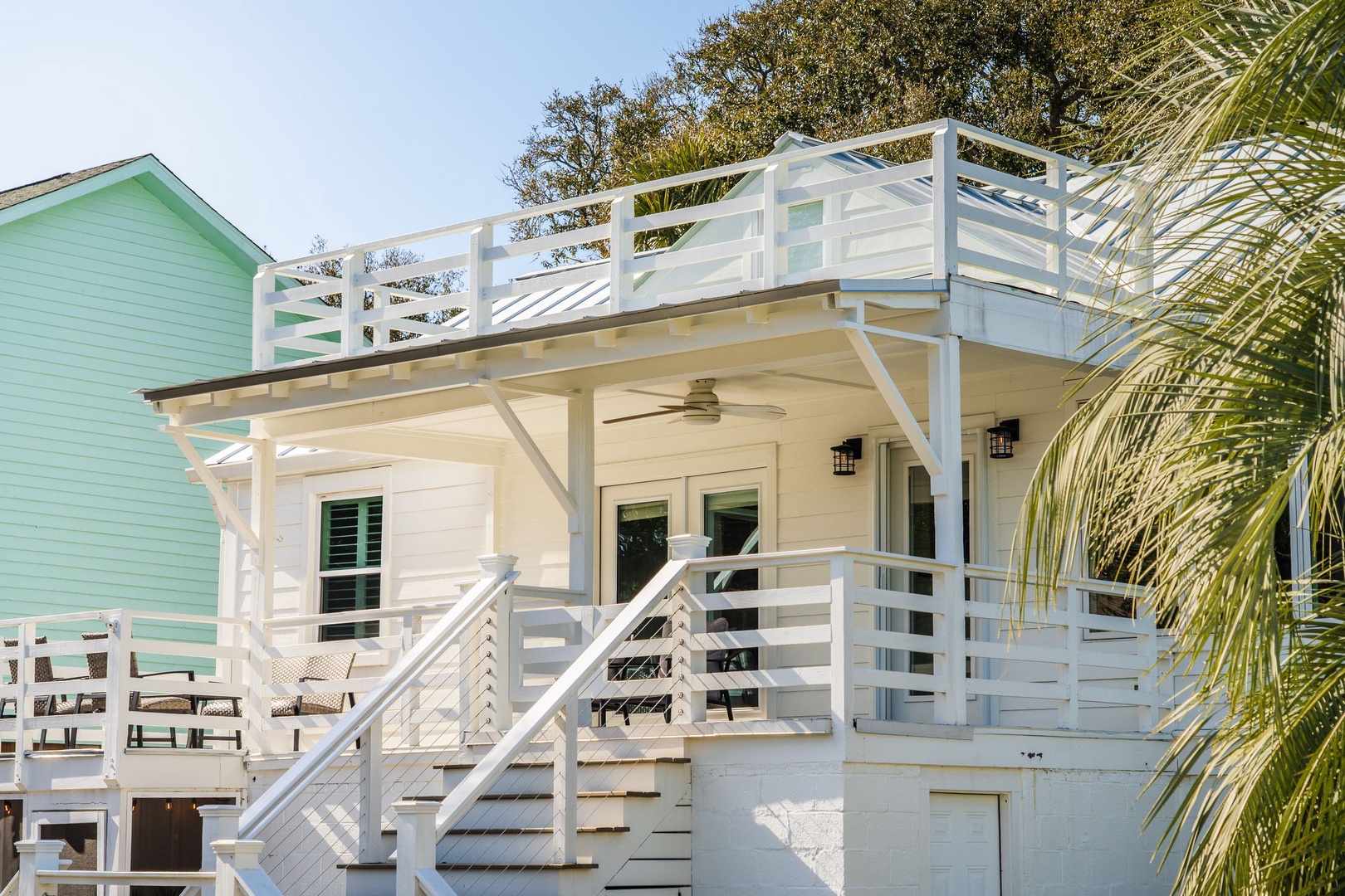 Tropical beach house featuring covered porch with ceiling fan, rooftop deck, and charming coastal architecture surrounded by lush palms.