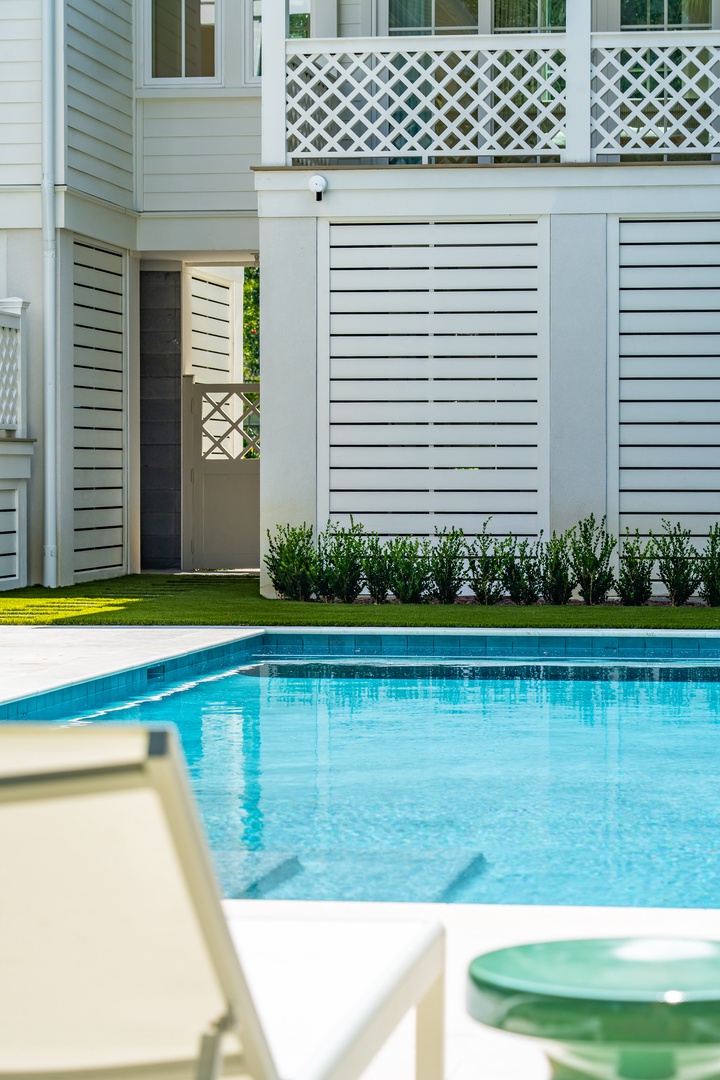 A bright, elegant property featuring a sparkling pool surrounded by lush lawn, classic white architecture, and inviting upper-level balcony with lattice detailing.