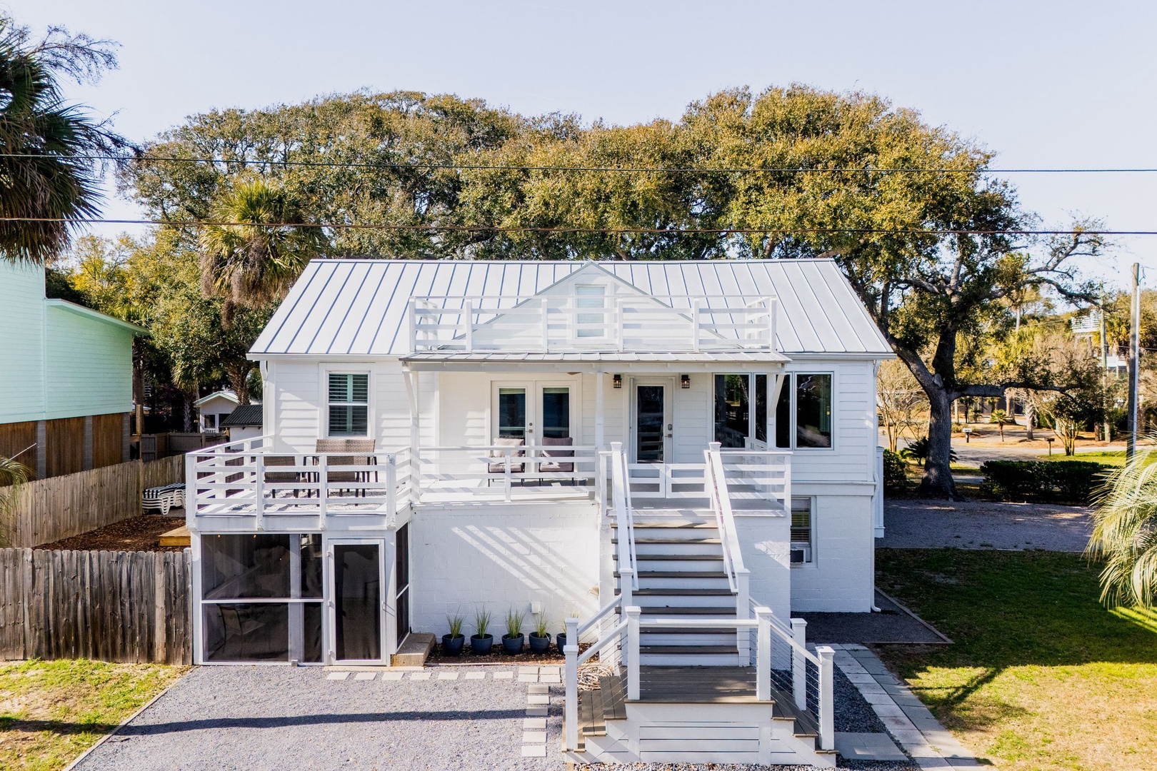Charming coastal cottage with white railings and metal roof, nestled among mature oak trees in a peaceful neighborhood setting.