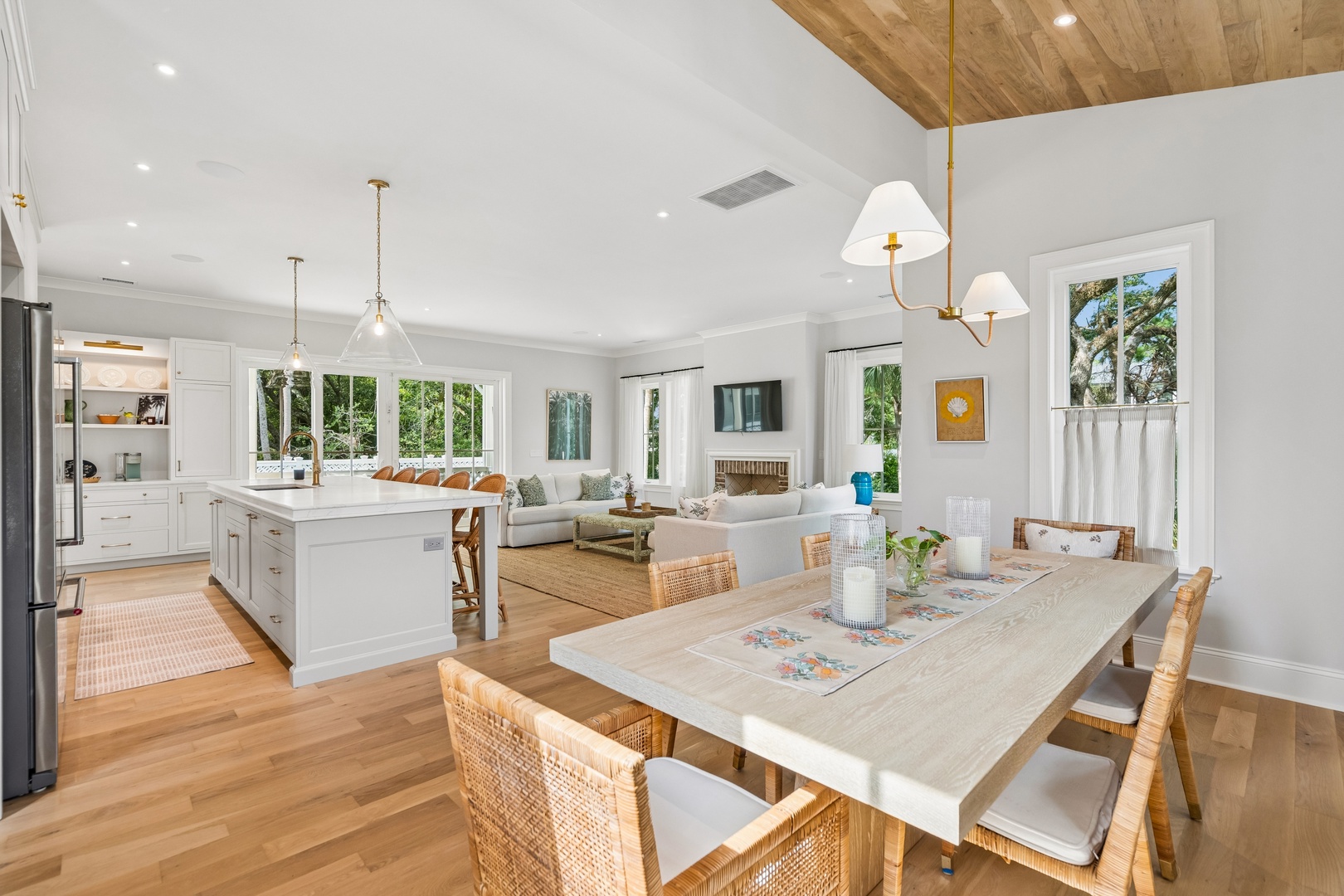 Your open-concept living space flows seamlessly from dining to kitchen to lounge, with natural light pouring through oversized windows. The vaulted wood ceiling, cozy fireplace, and woven accents create a warm coastal retreat perfect for gathering.