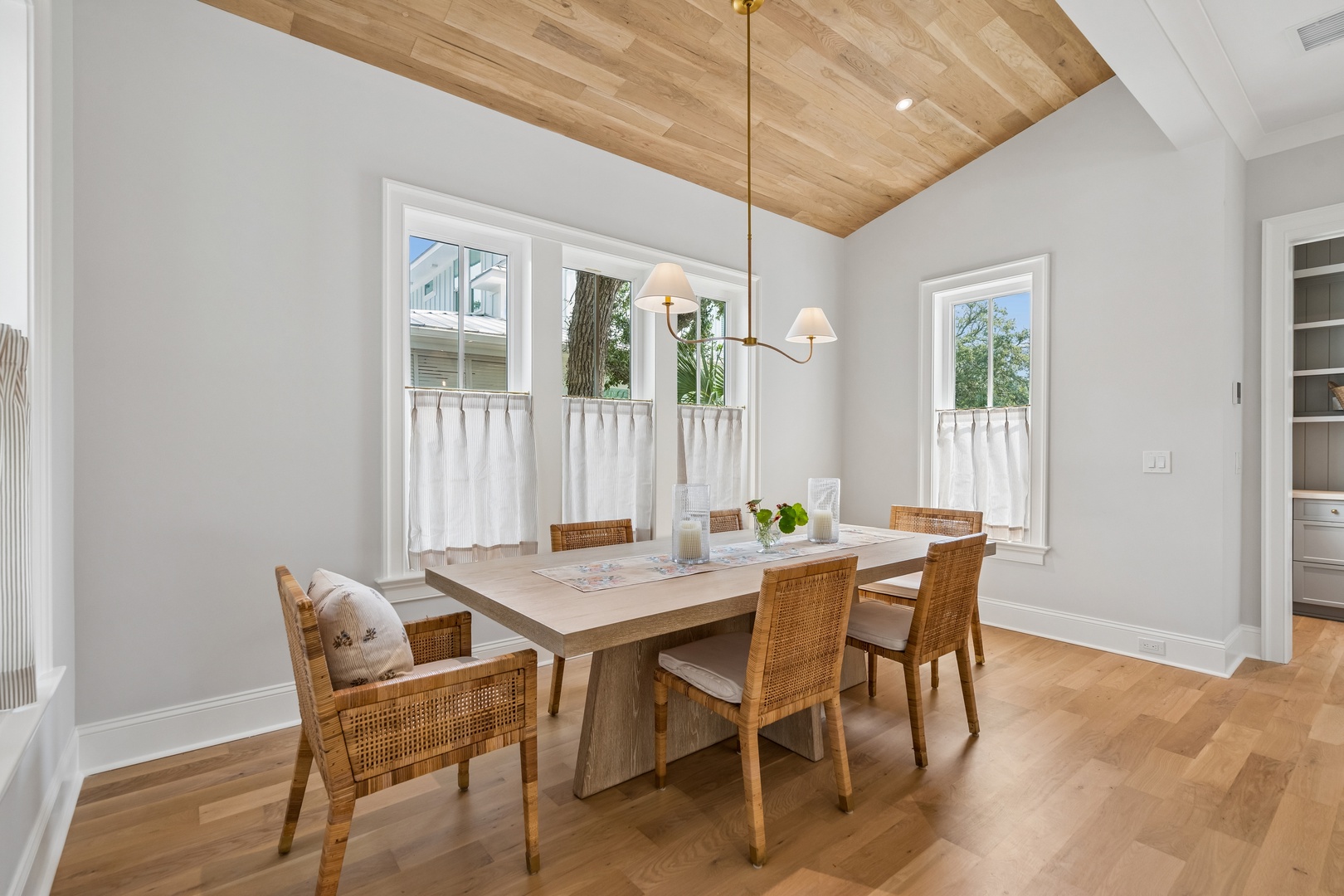 Gather around this beautiful dining table beneath the warm wood-toned ceiling, where natural light streams through charming café-curtained windows, perfect for your morning coffee or evening meals together.