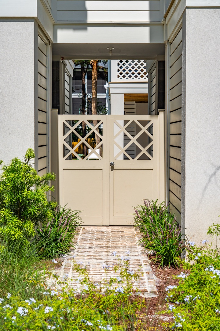 A charming lattice gate welcomes guests to this coastal property, framed by lush greenery and flowering plants along a brick pathway.