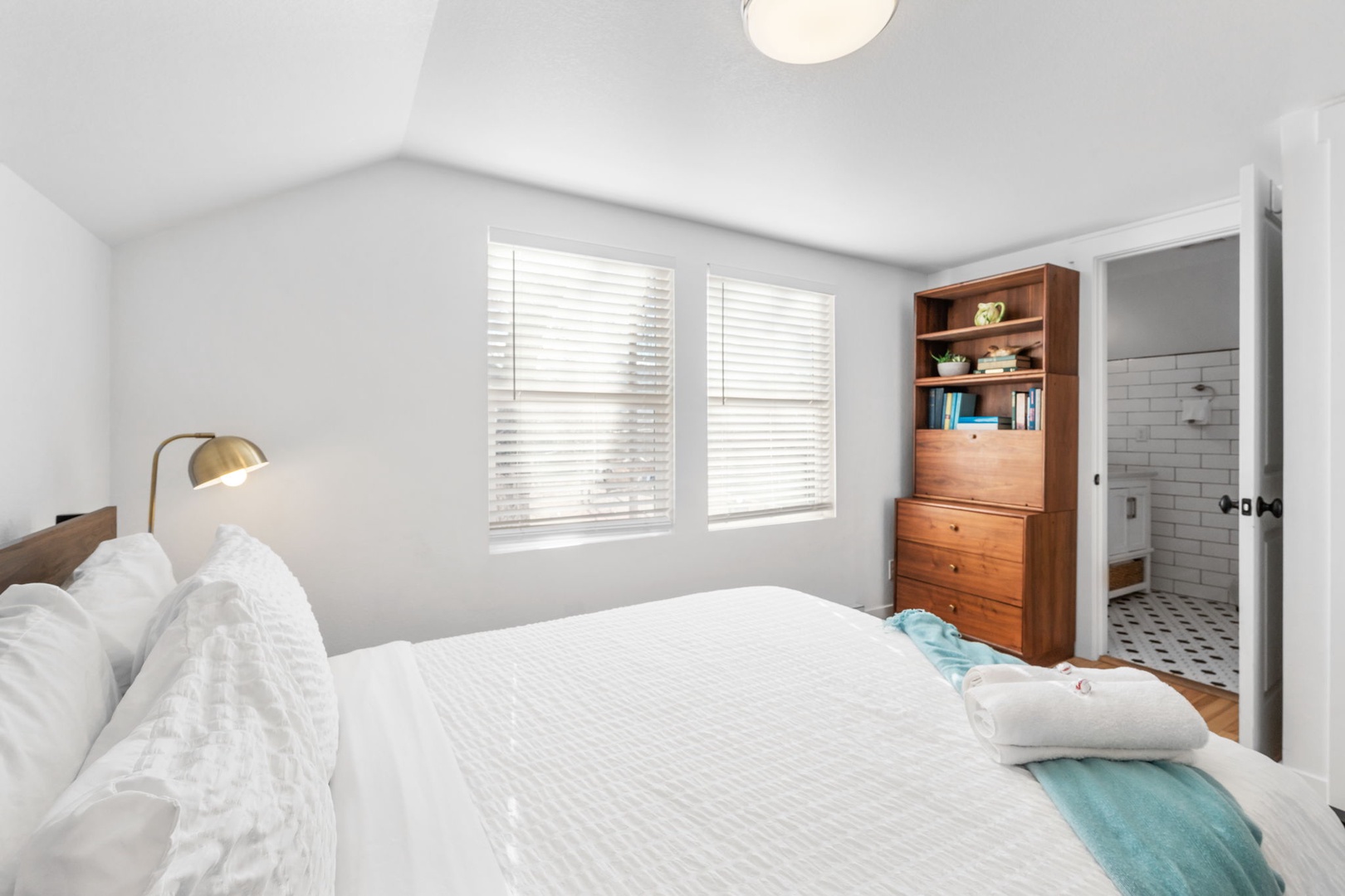 Rest in this bright bedroom where natural light filters through blinds, creating a peaceful retreat with warm wood accents and an en-suite bathroom nearby.