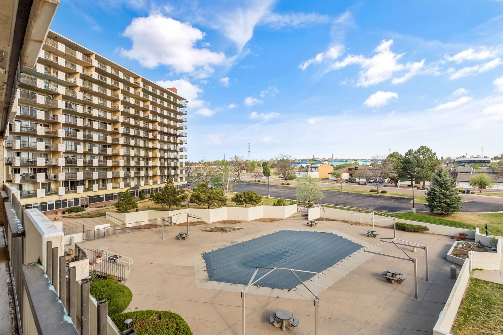 Multi-story property building with covered pool area, landscaped grounds, and nearby parking visible under bright blue skies and scattered clouds.