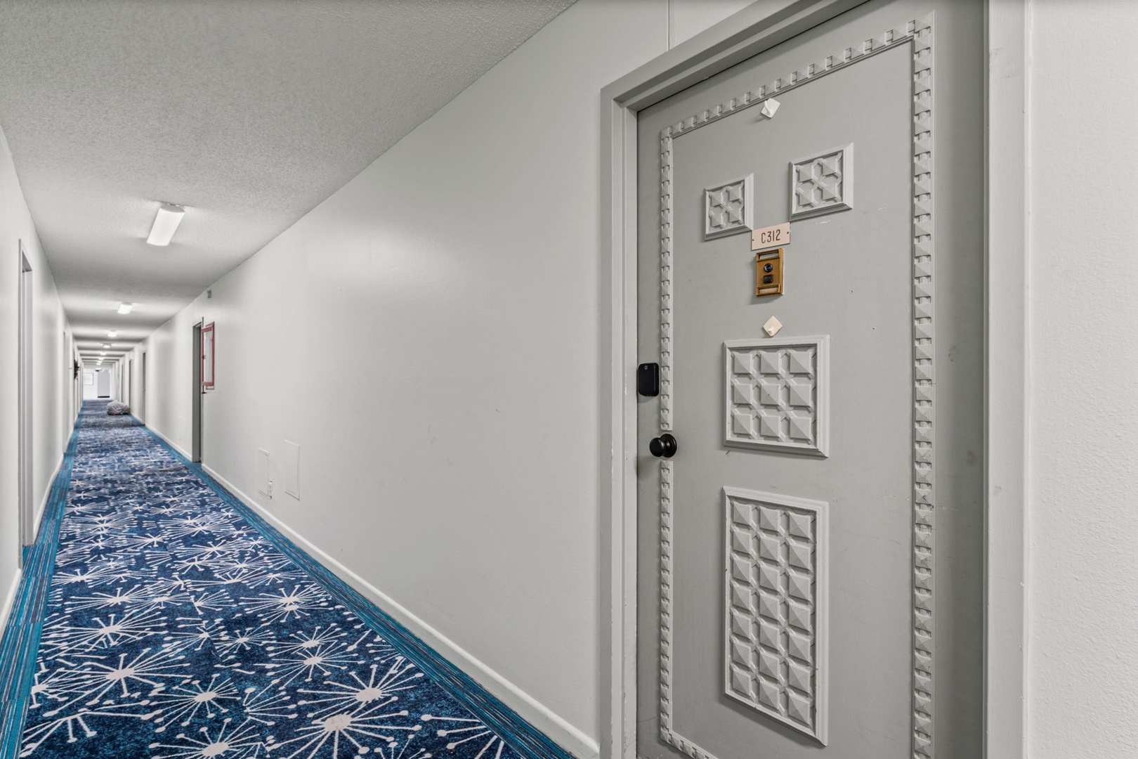 The property features bright, well-lit hallways with modern carpeting and clean white walls, leading you comfortably to your room entrance with a secure keyless entry system.