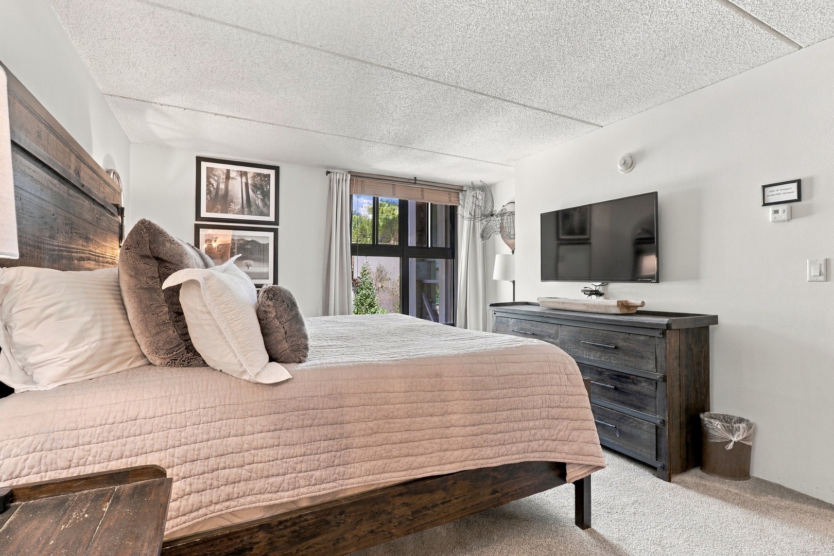Sink into plush bedding and unwind in your serene retreat. Warm wood tones and soft lighting create a cozy atmosphere, while natural light from the window brightens your space beautifully.