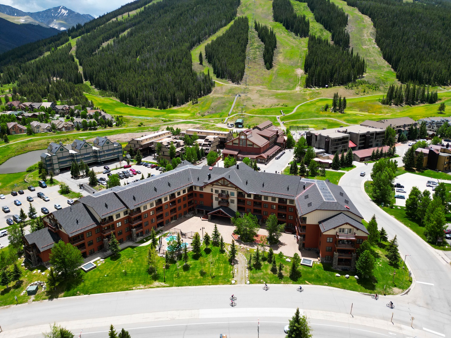 Aerial view of a mountain resort village nestled at the base of forested ski slopes, surrounded by green summer meadows and dramatic alpine peaks.