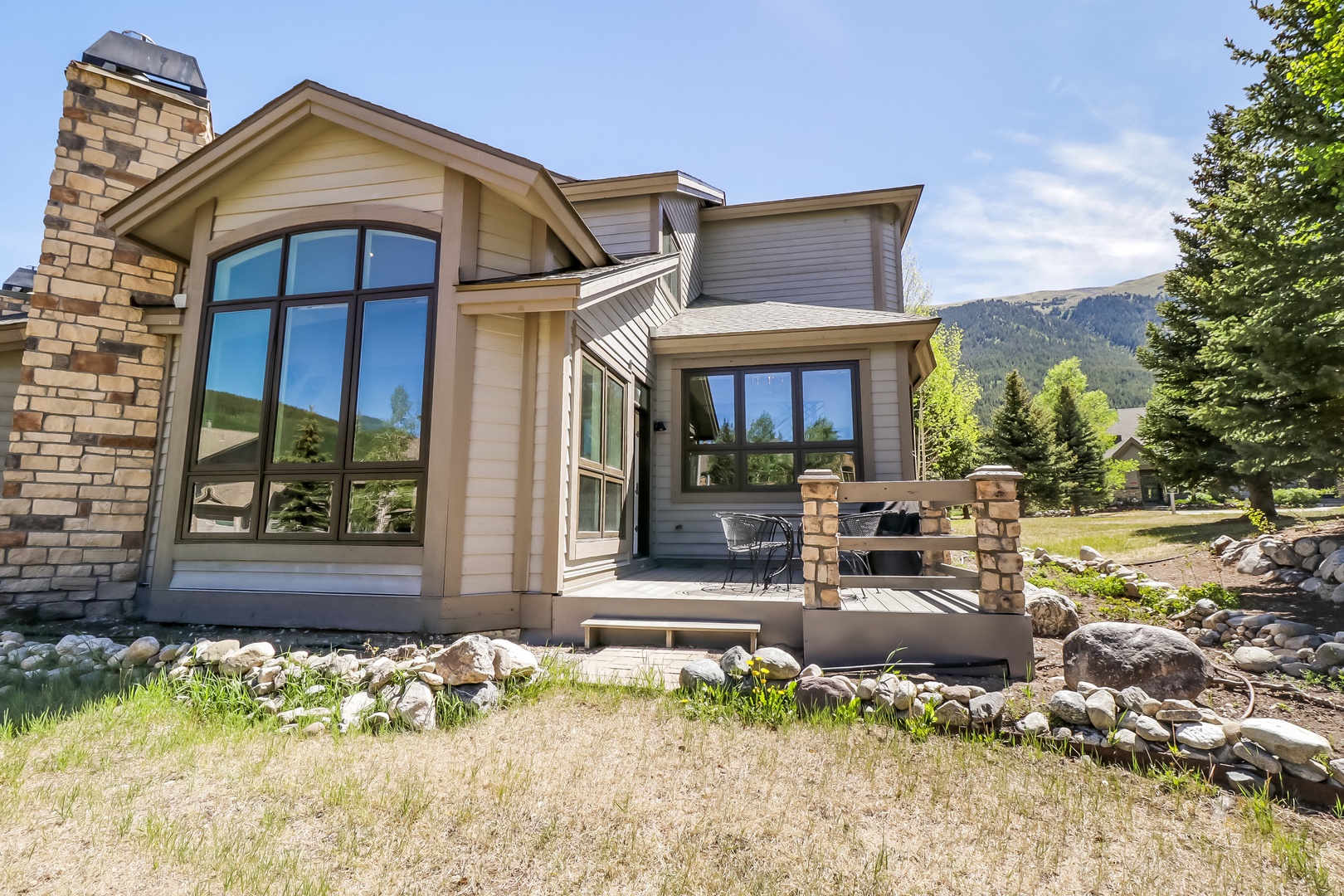 A charming mountain retreat featuring stone accents, expansive windows, and a welcoming patio surrounded by natural landscaping and stunning mountain vistas.