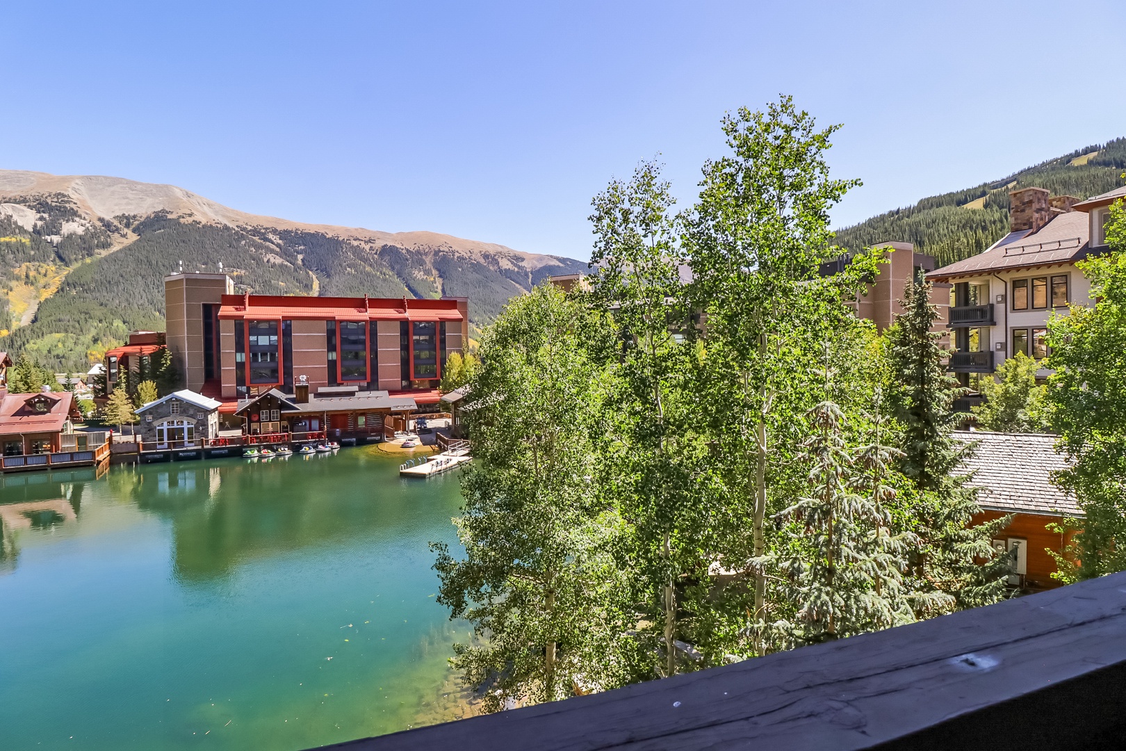A lakeside alpine village framed by towering mountains and forested slopes. The turquoise waters and surrounding buildings showcase the charming mountain-town atmosphere of the area.