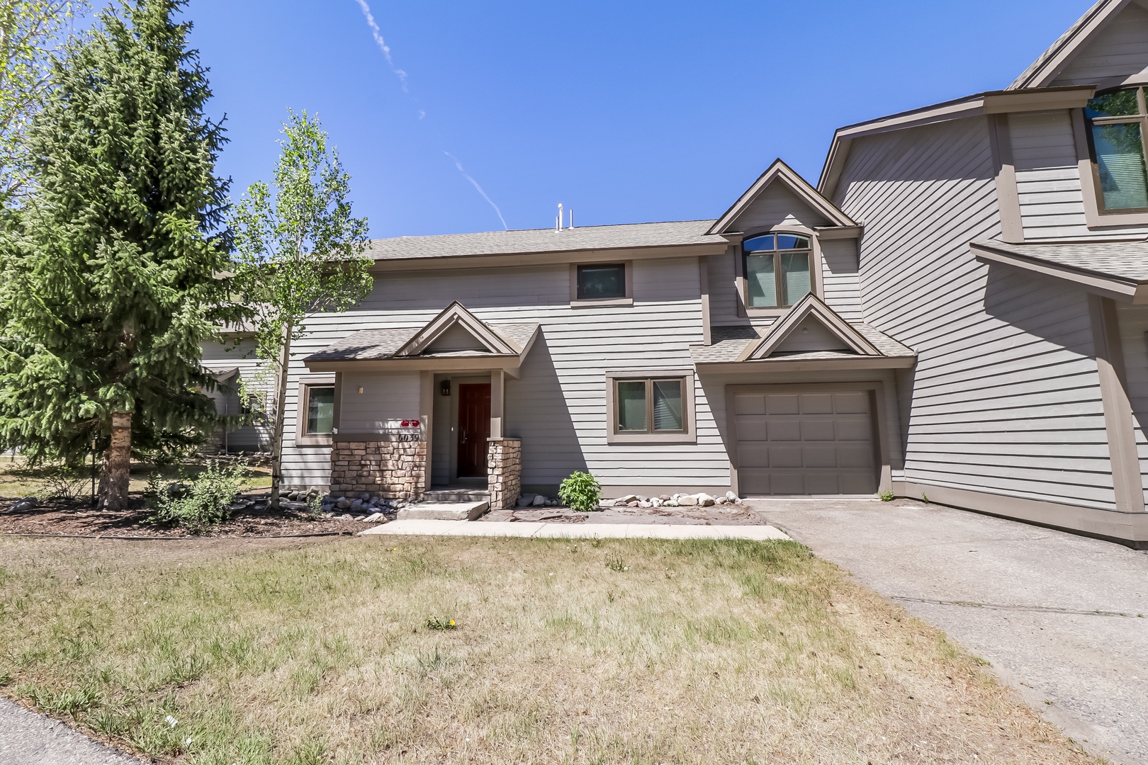 A welcoming two-story vacation rental with stone accents, attached garage, and mountain-area charm framed by mature trees and sunny skies.