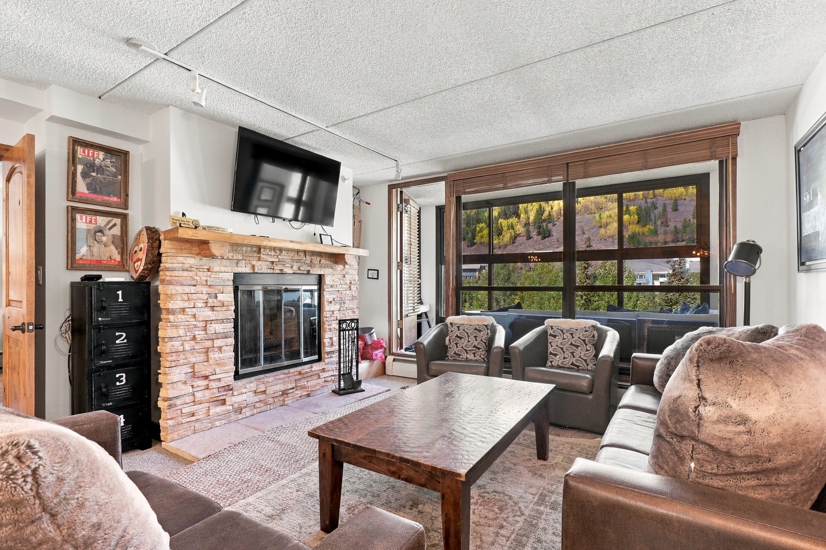 Step into a welcoming living room where rustic charm meets mountain comfort. Settle into plush seating before a warm brick fireplace, and let your gaze drift through the large windows to stunning mountain and greenery views just beyond.