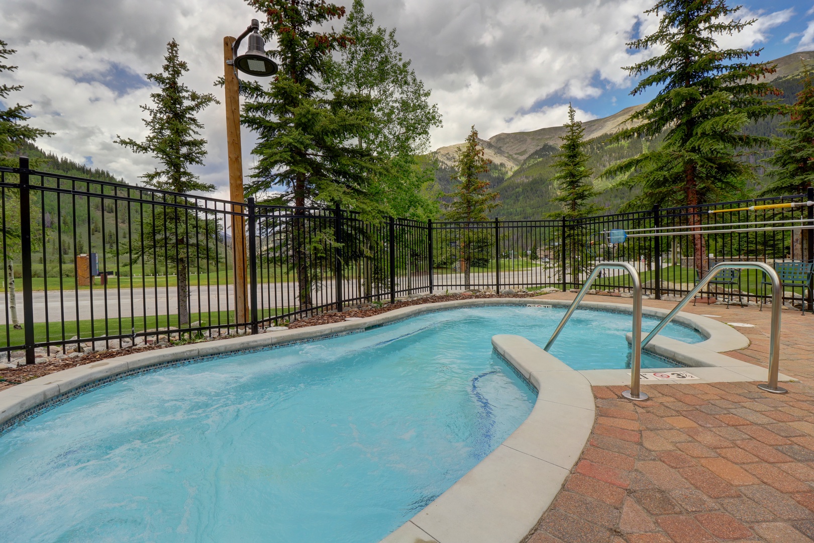 An outdoor pool and hot tub set against towering mountain peaks and evergreen forests. Perfect for relaxing after a day of mountain adventures.