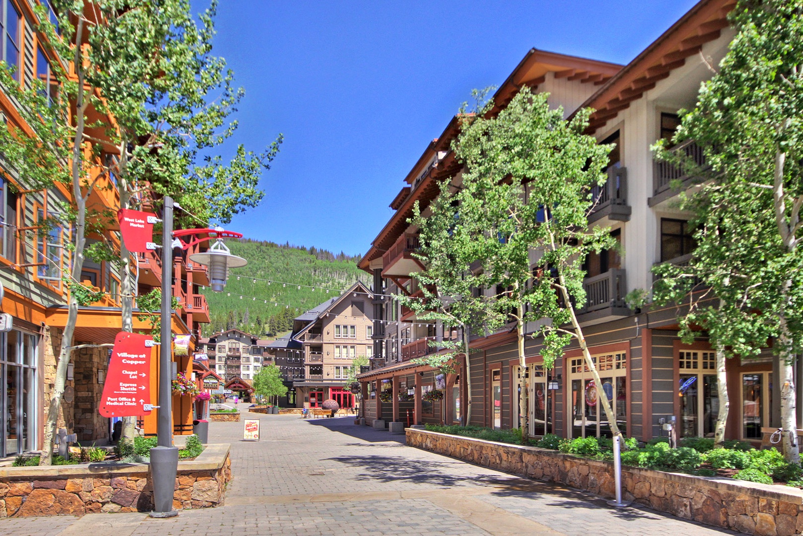 A charming pedestrian walkway winds through the village center, lined with shops and restaurants, with mountain vistas framing the horizon beyond tree-lined pathways and alpine-style architecture.