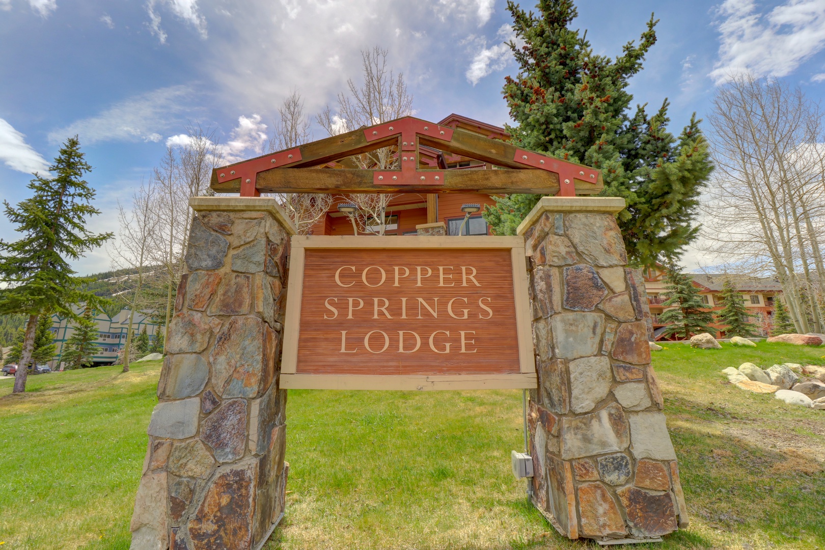 The property entrance features a distinctive stone and timber monument sign identifying the location within a mountain setting surrounded by evergreens and natural landscape.