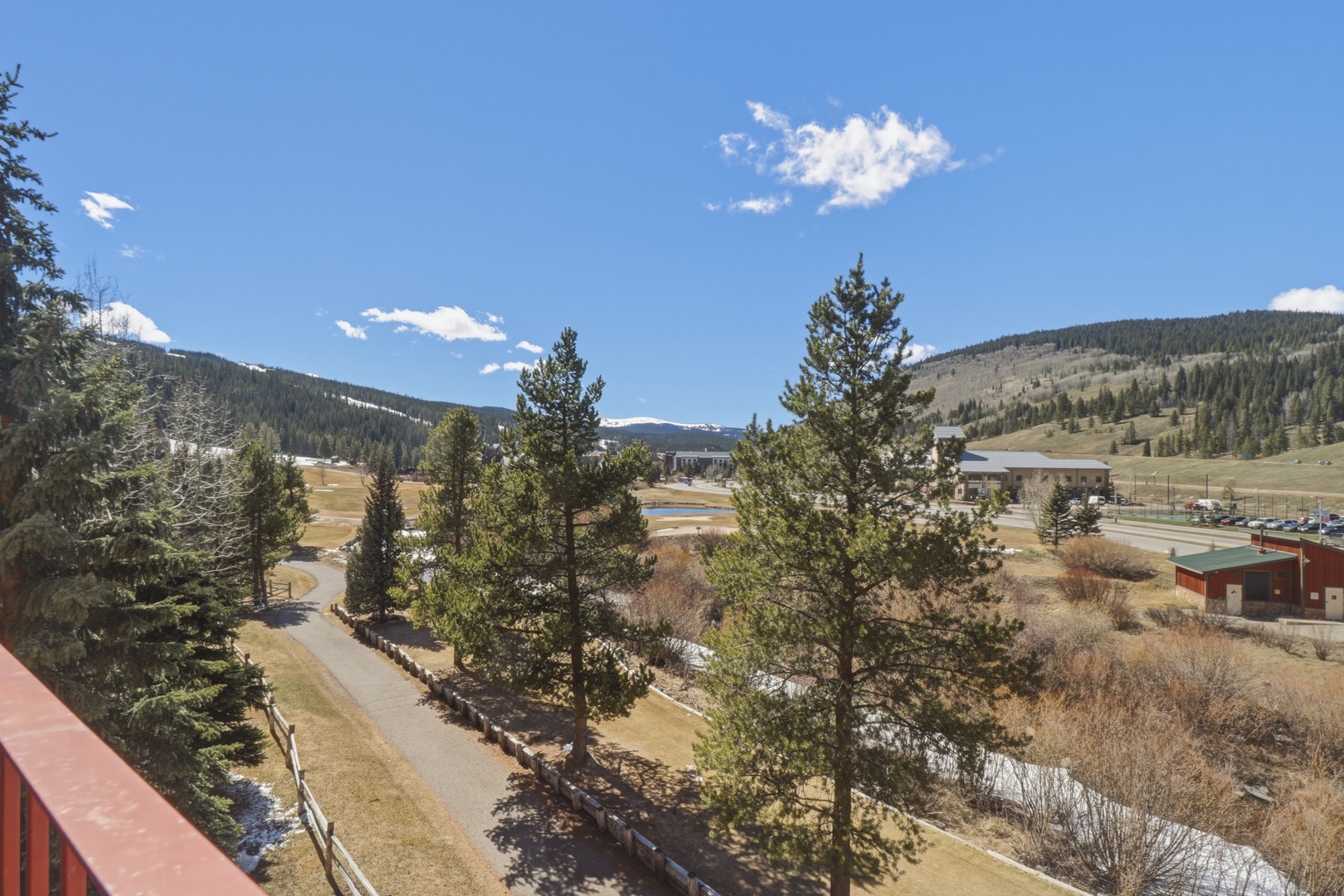 Stunning mountain valley panorama spreads before this elevated terrace, featuring rolling hills, evergreen forests, and distant peaks under clear blue skies.