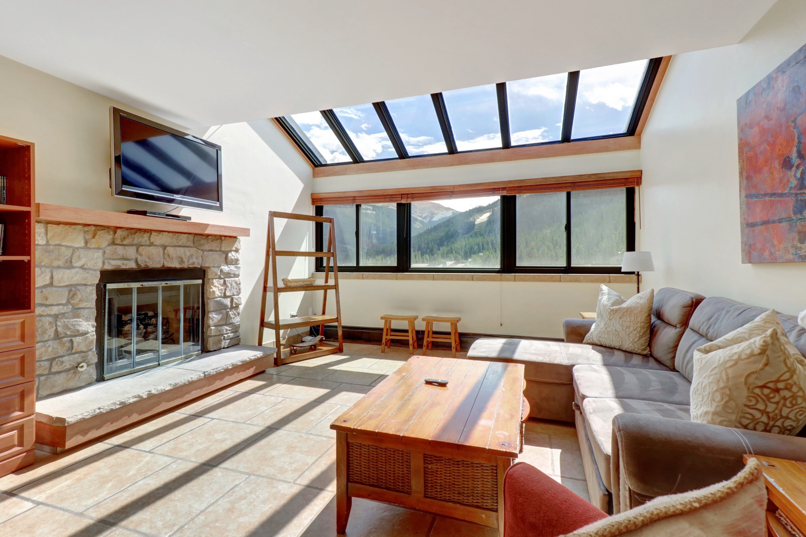 Bright and airy living room featuring a large skylight and expansive windows that showcase breathtaking mountain scenery. Cozy stone fireplace and comfortable sectional sofa make it a perfect retreat after a day on the slopes.