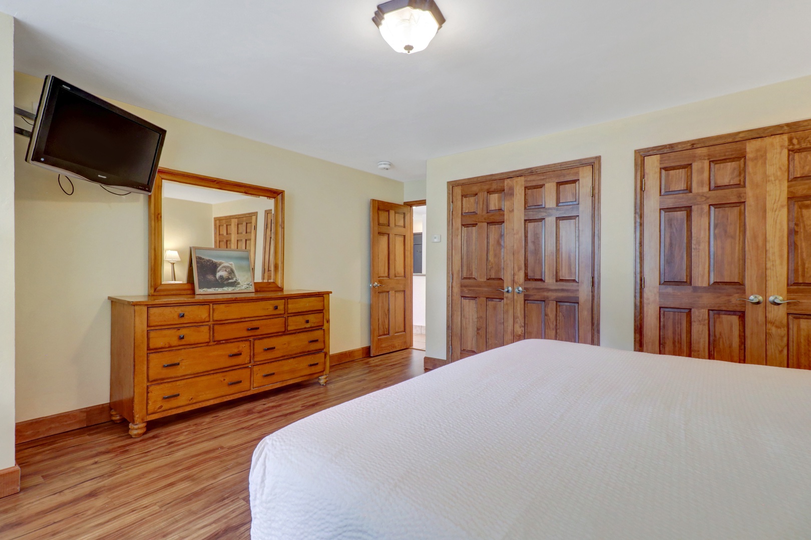 Comfortable bedroom featuring rich wood furnishings, a large dresser with mirror, and generous closet space. Includes a wall-mounted TV for convenient in-room entertainment.
