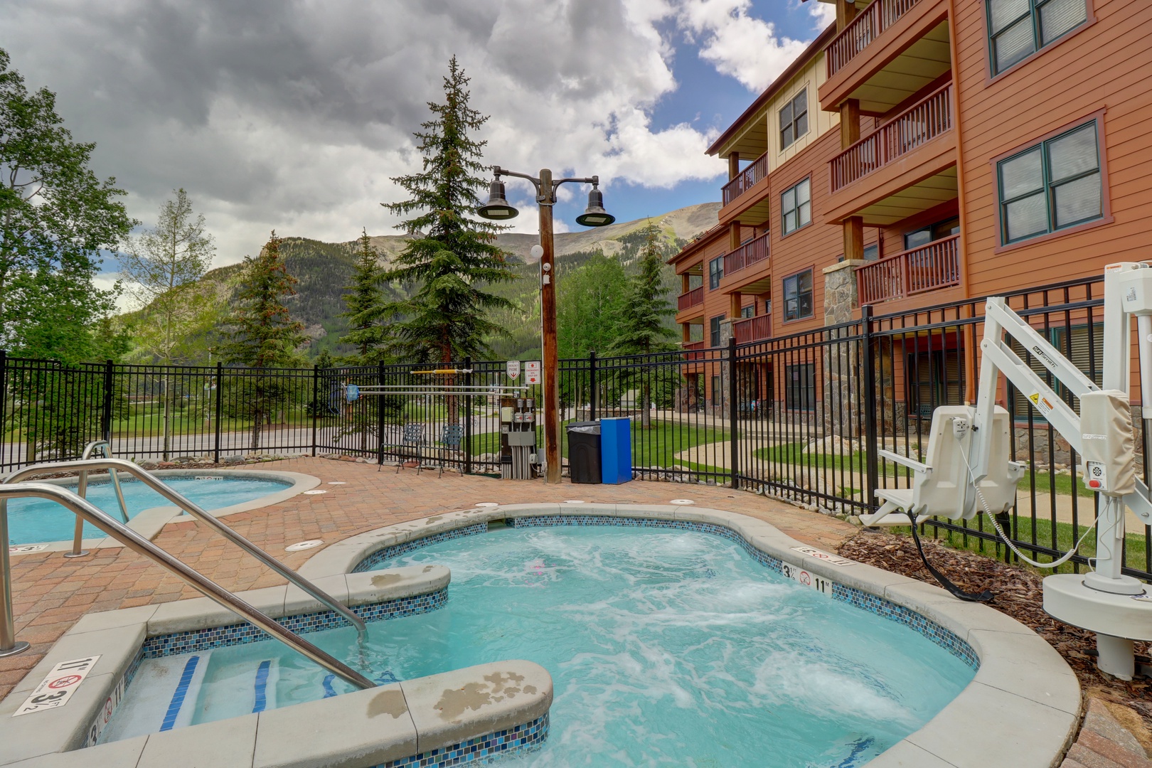 A welcoming mountain resort featuring hot tubs, pool, and multi-story lodgings with balconies, surrounded by evergreen trees and dramatic alpine peaks under partly cloudy skies.
