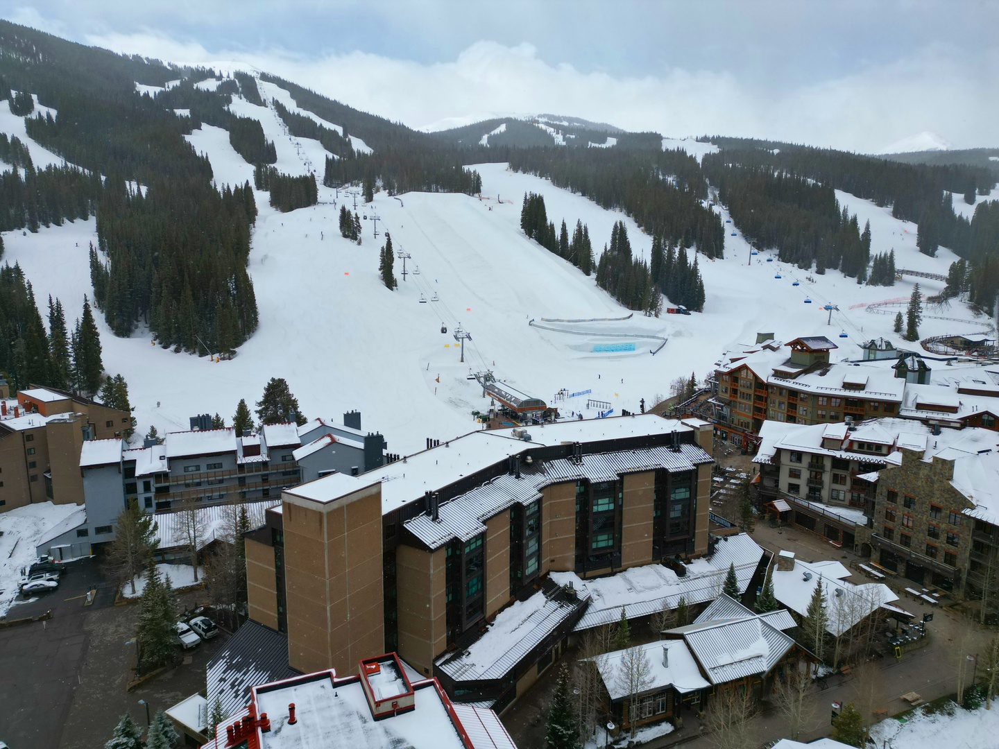 An aerial view captures a mountain village nestled at the base of snowy ski slopes, showcasing ski lifts and winter terrain in the surrounding area.