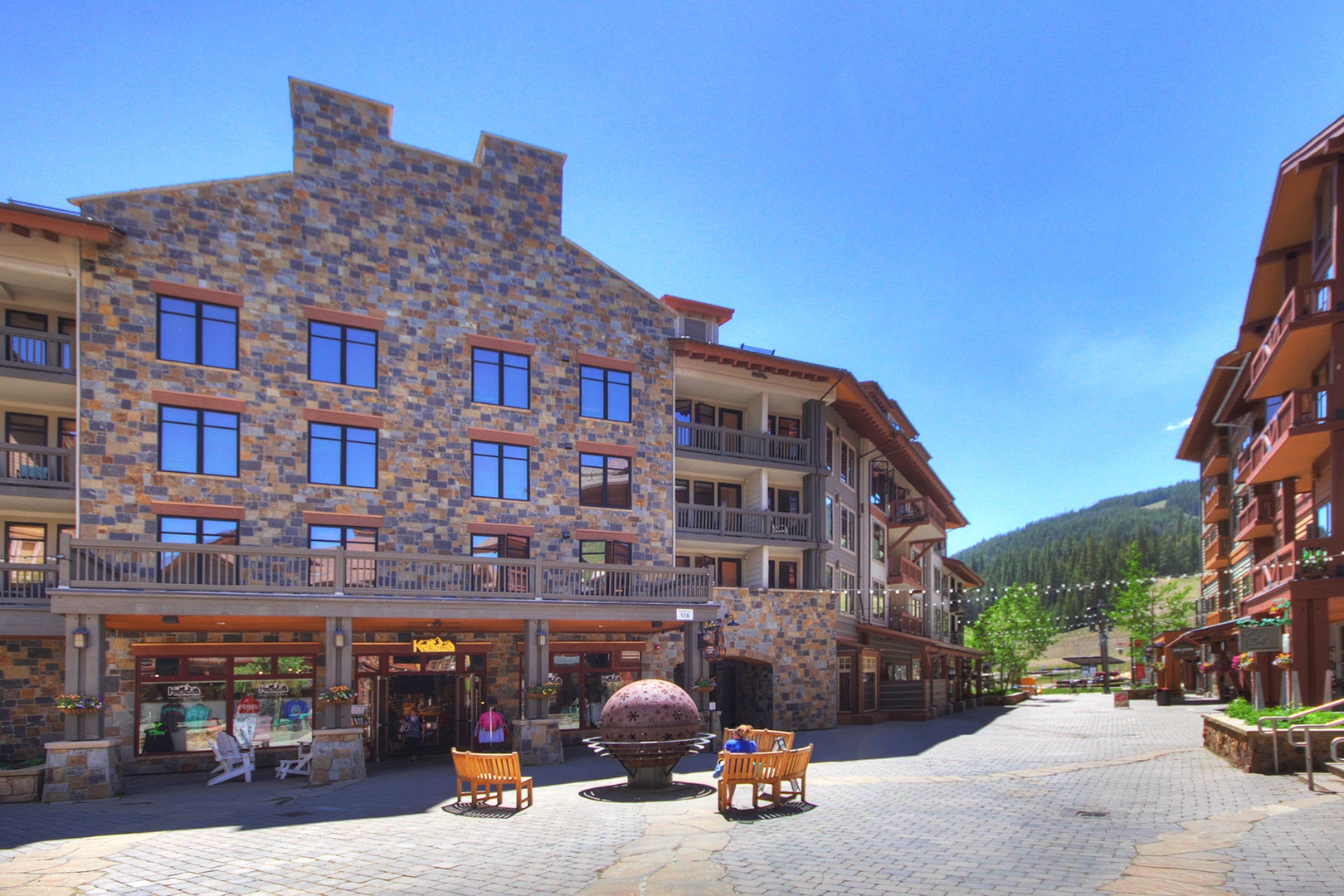 The property is located within a charming alpine village setting, featuring stone-clad buildings and pedestrian-friendly walkways surrounded by forested mountain slopes.
