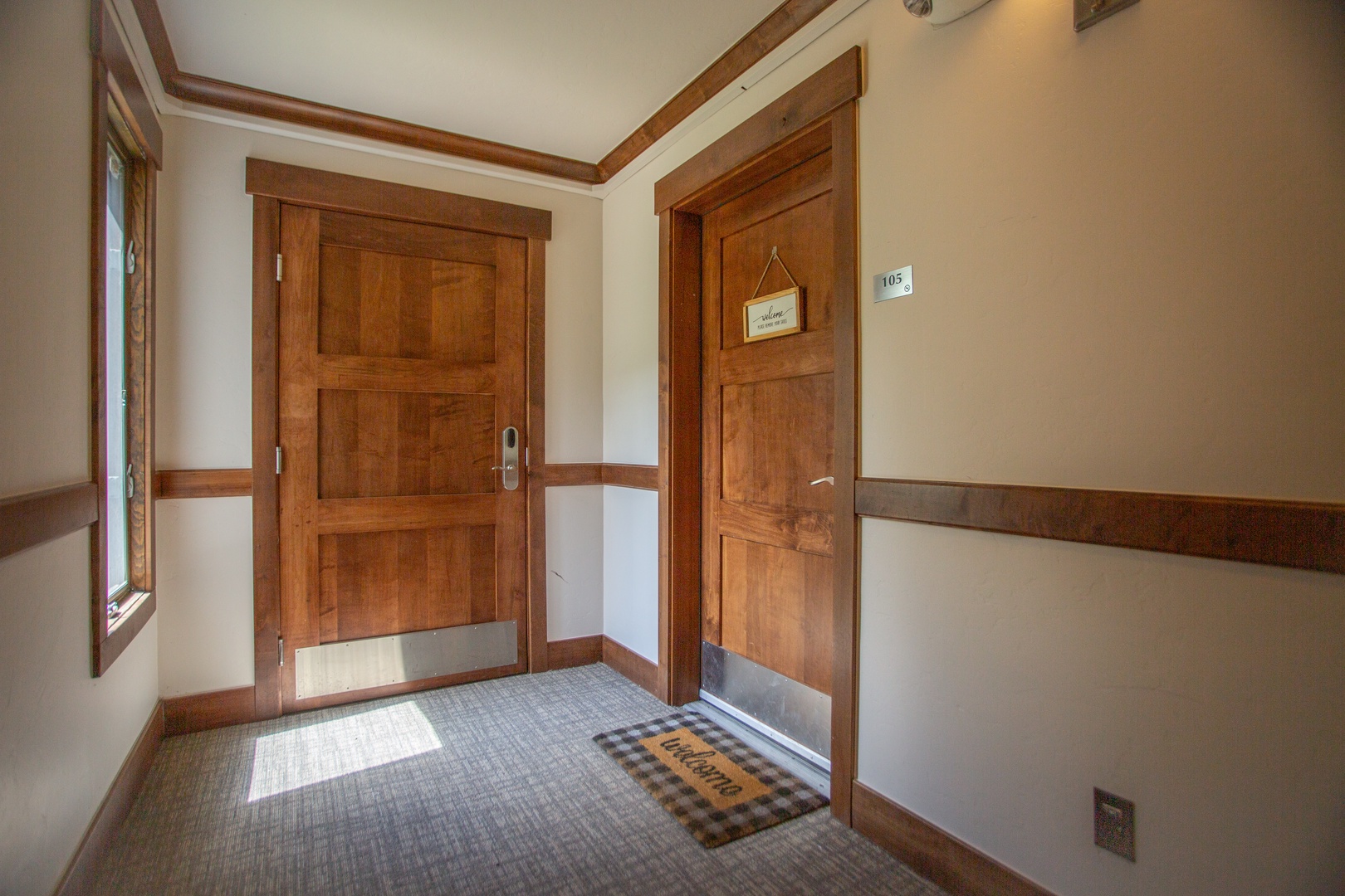The hallway leading to your room invites you in with warm wooden accents and natural light streaming through the window. You'll find your private retreat just beyond the charming welcome mat—ready for your arrival.