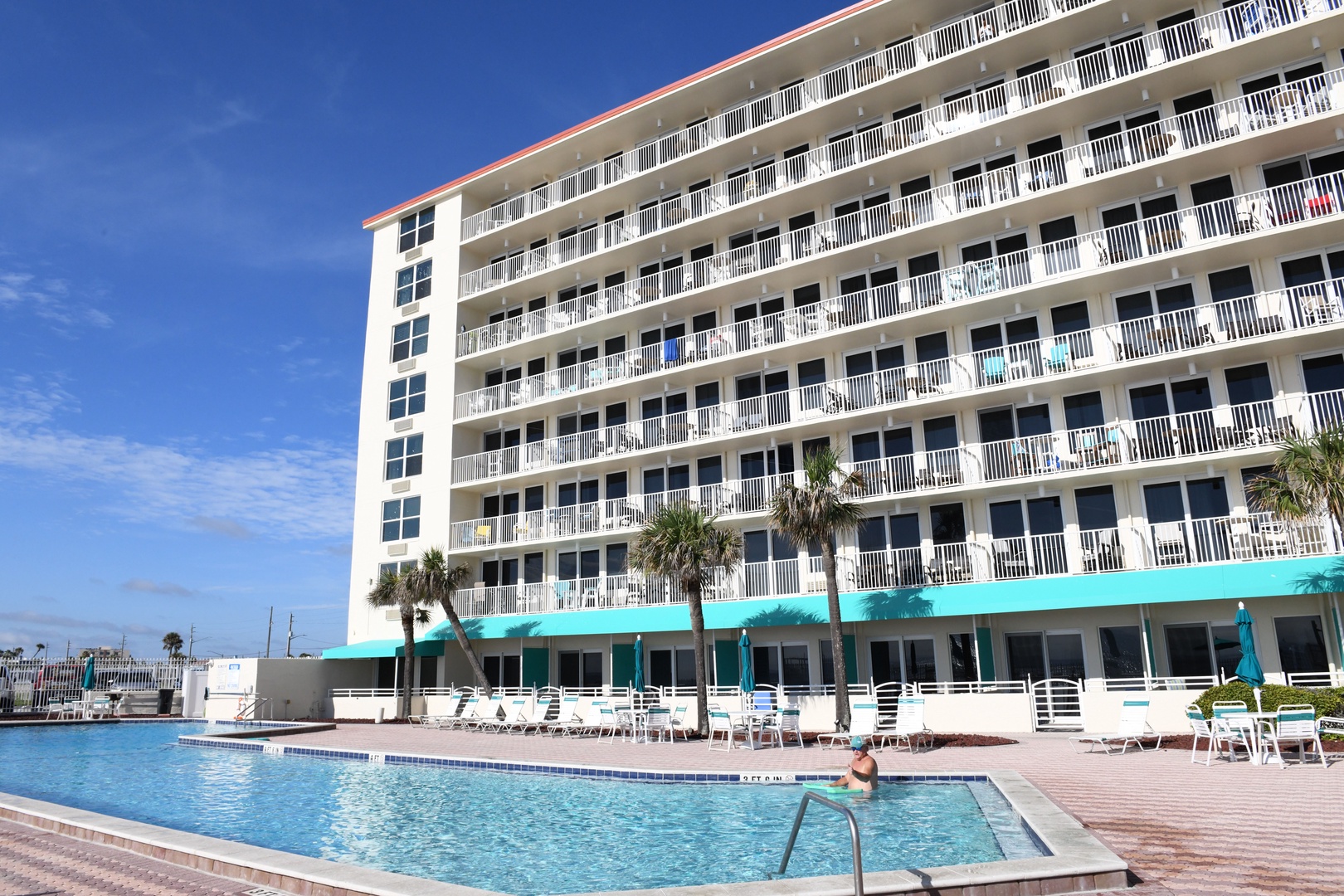 A multi-story beachside property featuring a sparkling outdoor pool, sun loungers, and palm trees against a clear blue sky—ideal for relaxing vacation days.