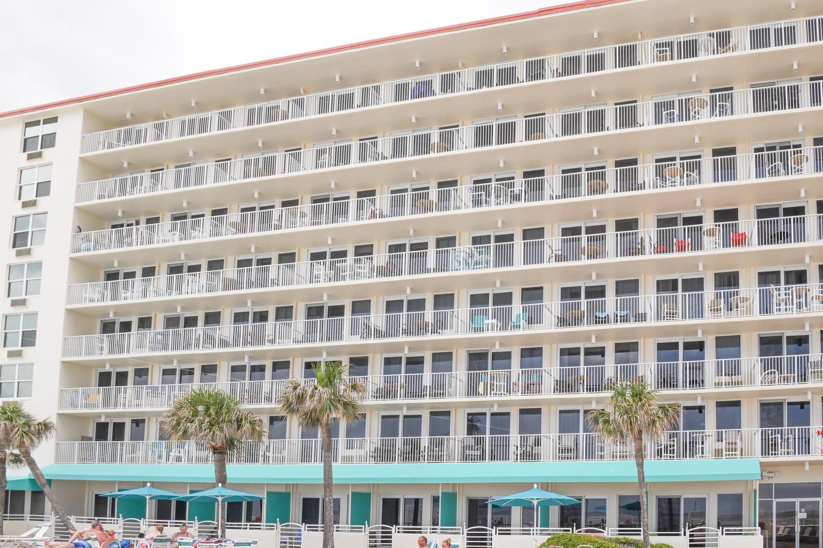 Beachfront property featuring multiple floors of balconies and ground-level access to the sandy shore, surrounded by swaying palms.