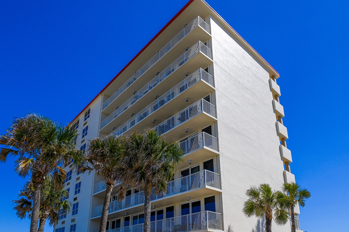 Modern beachfront building with private balconies surrounded by tropical palms under clear blue skies.