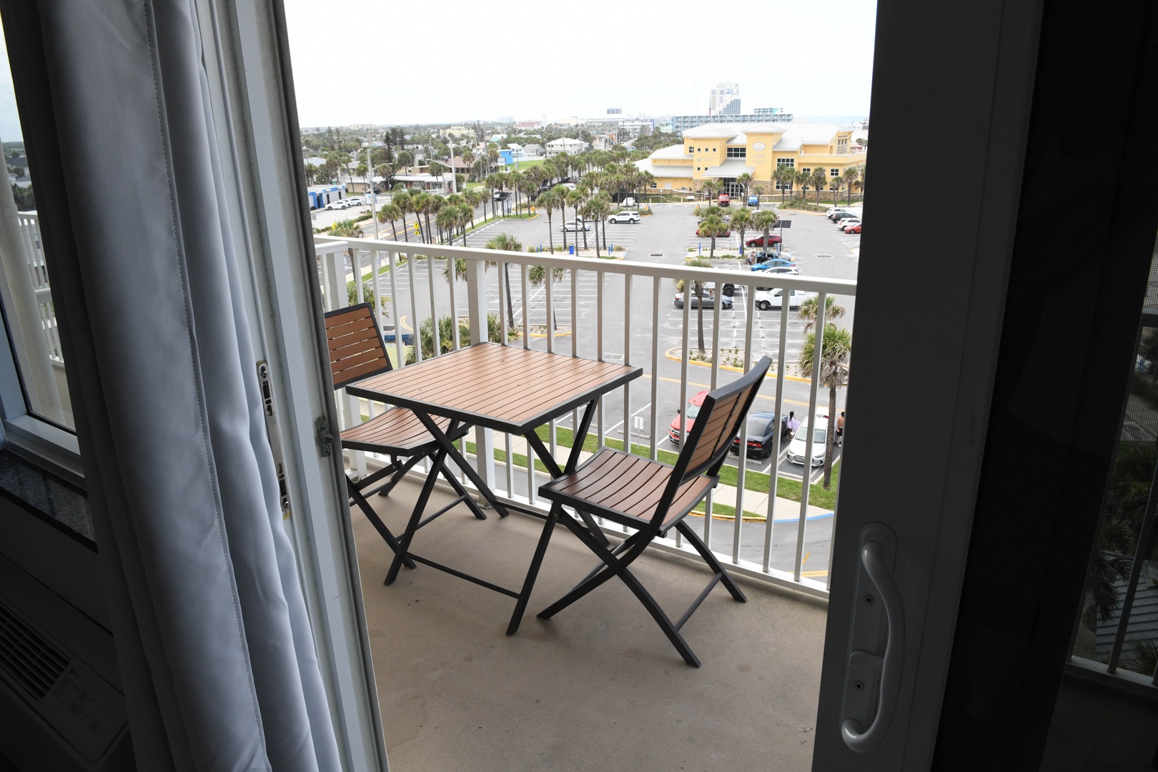 Balcony overlooks a palm-lined plaza and surrounding cityscape, offering an elevated vantage point with outdoor seating for observing the area's shops and parking below.