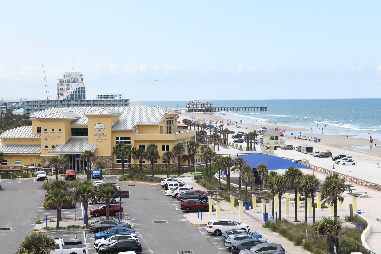 Coastal beach scene with sandy shore, palm trees, nearby buildings, and ocean view. Parking area provides convenient beach access.