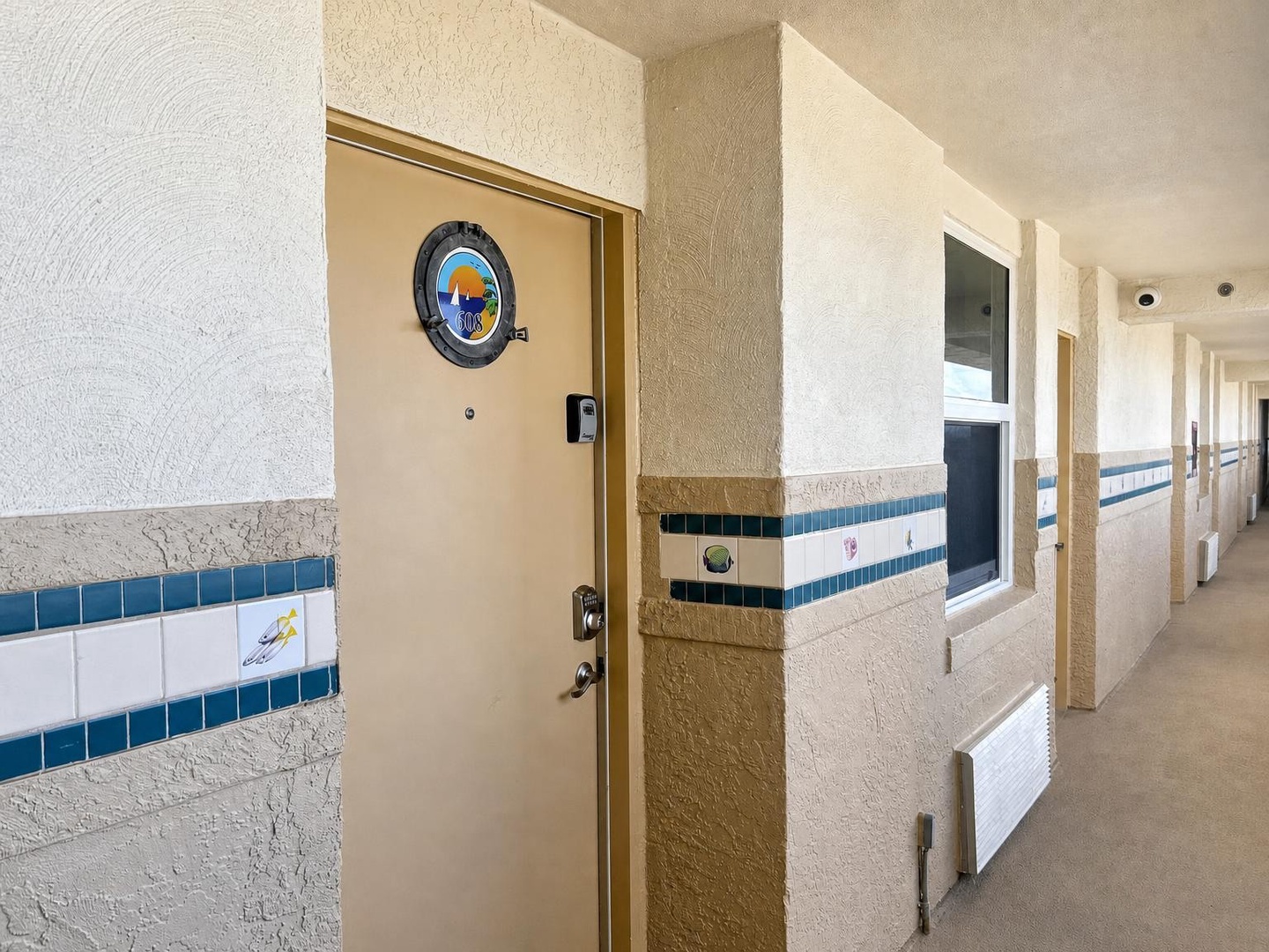 A sun-lit outdoor corridor leads to your vacation rental entrance, marked by cheerful tilework and a custom doorplate that captures the spirit of your seaside escape.