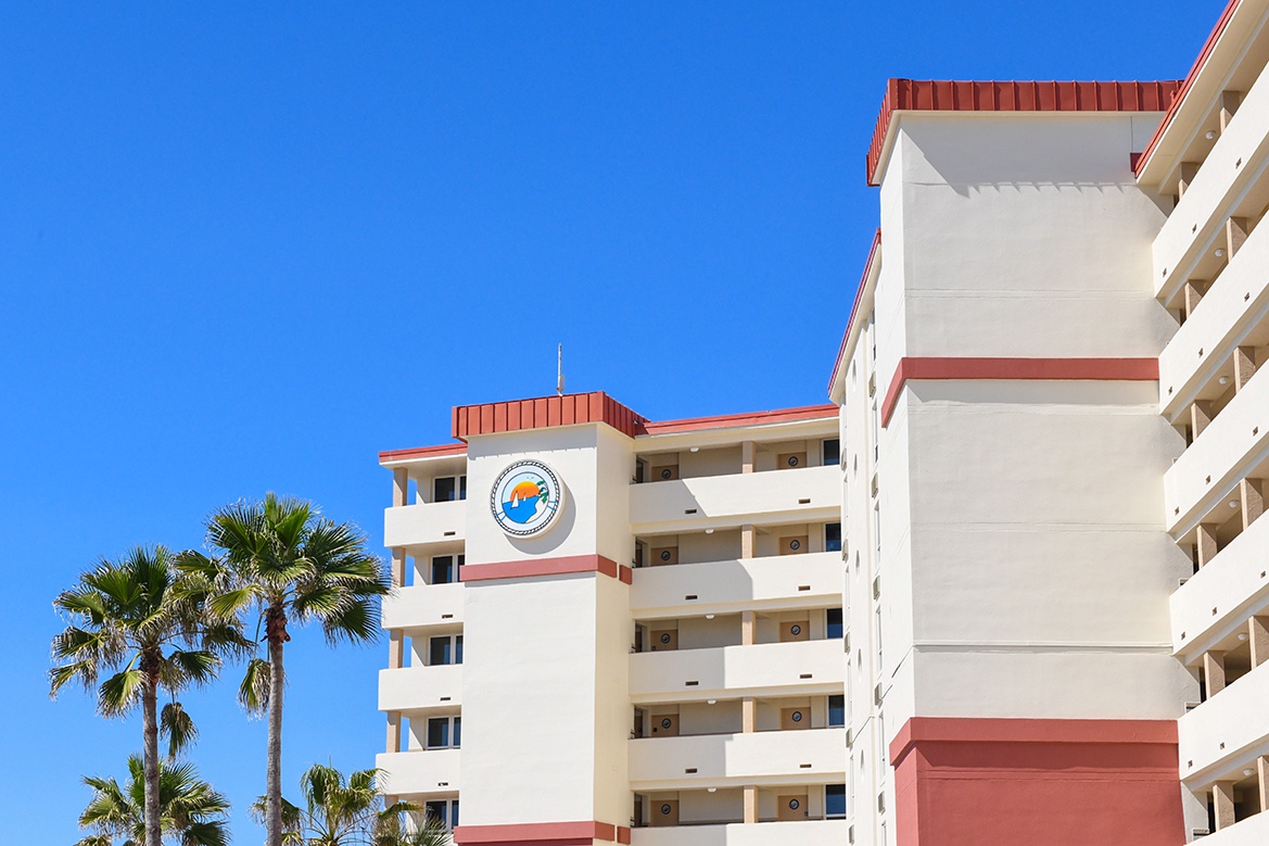 Multi-story property building with distinctive red roof accents and balconies under clear blue skies, surrounded by tropical palm trees.