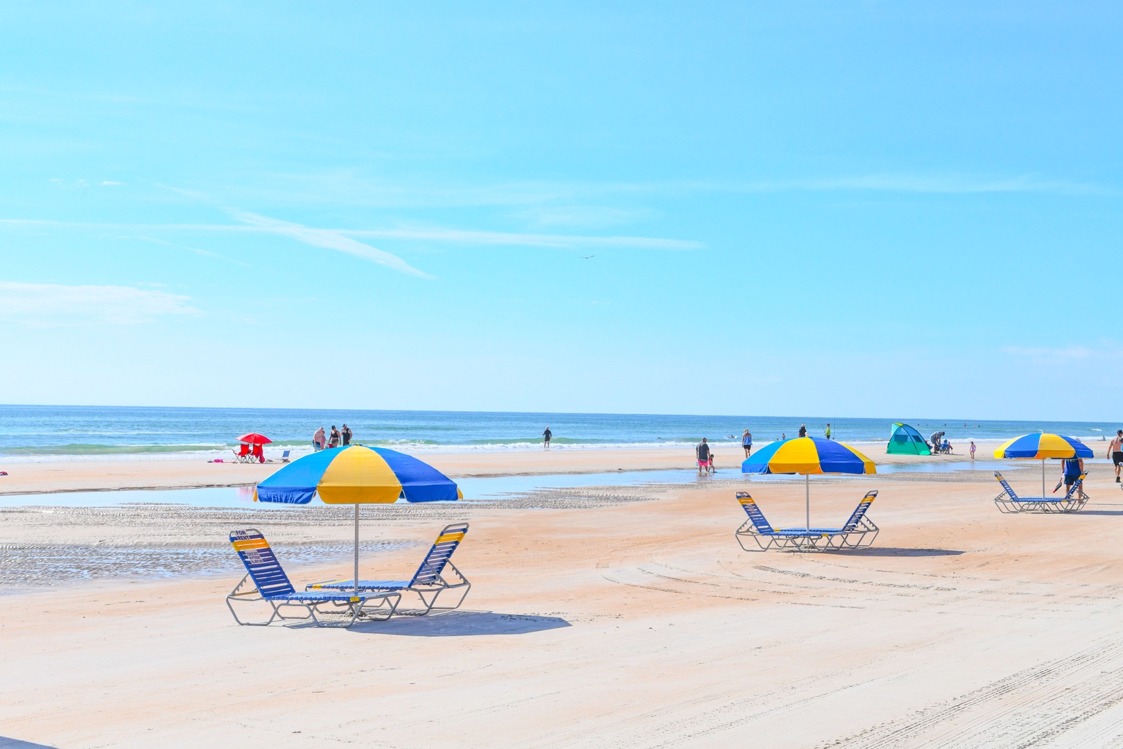 Sandy beach with colorful umbrellas and lounge chairs stretches along calm ocean waters under clear blue skies.