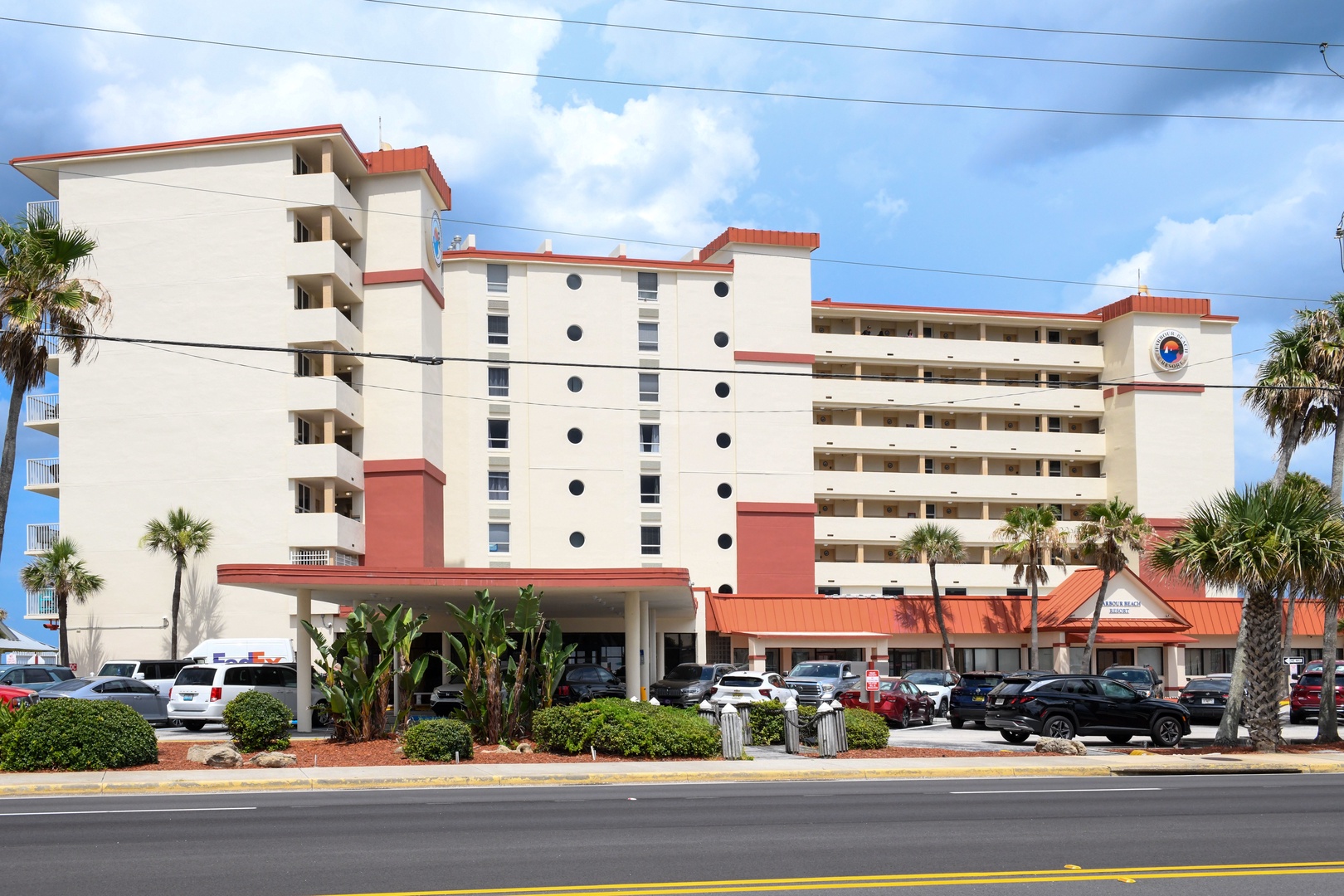 A multi-story beachfront property featuring white and coral architecture, surrounded by tropical palms and convenient parking, located in a coastal resort area.