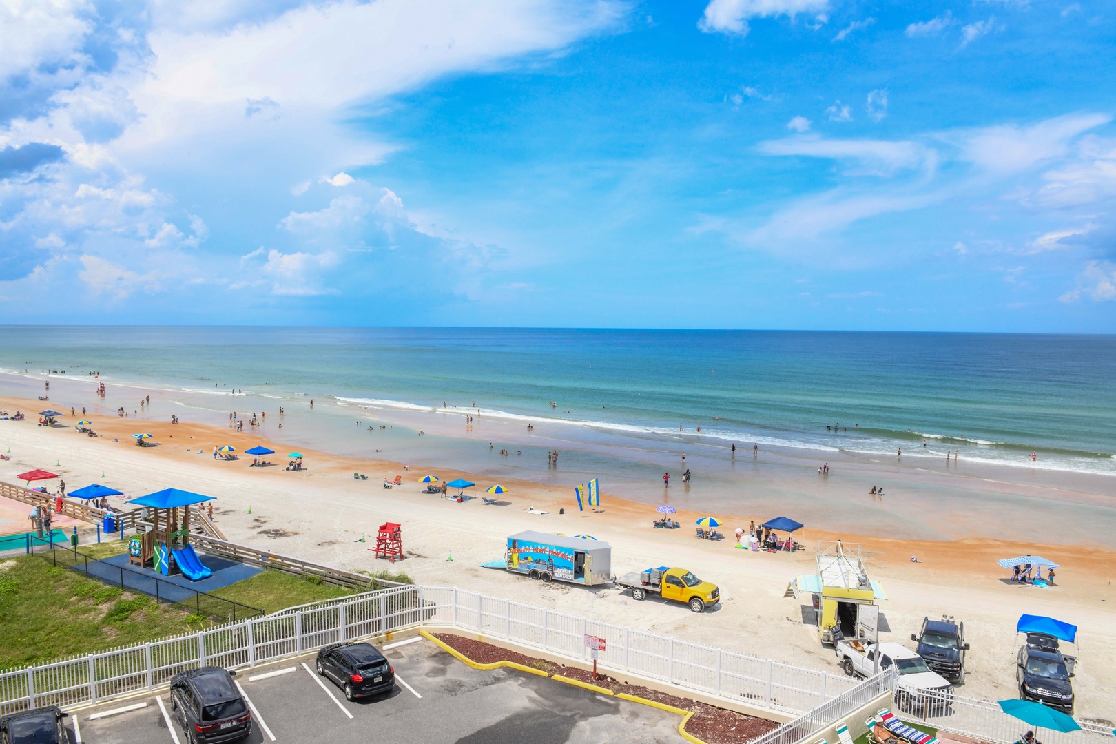 An aerial view capturing the property's prime beachfront location, with golden sand, vibrant umbrellas, gentle surf, and a colorful children's playground welcoming families to Florida's coast.