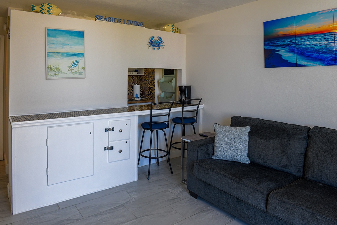 Your cozy kitchenette and living space blend coastal charm with modern comfort, featuring ocean artwork and comfy seating.