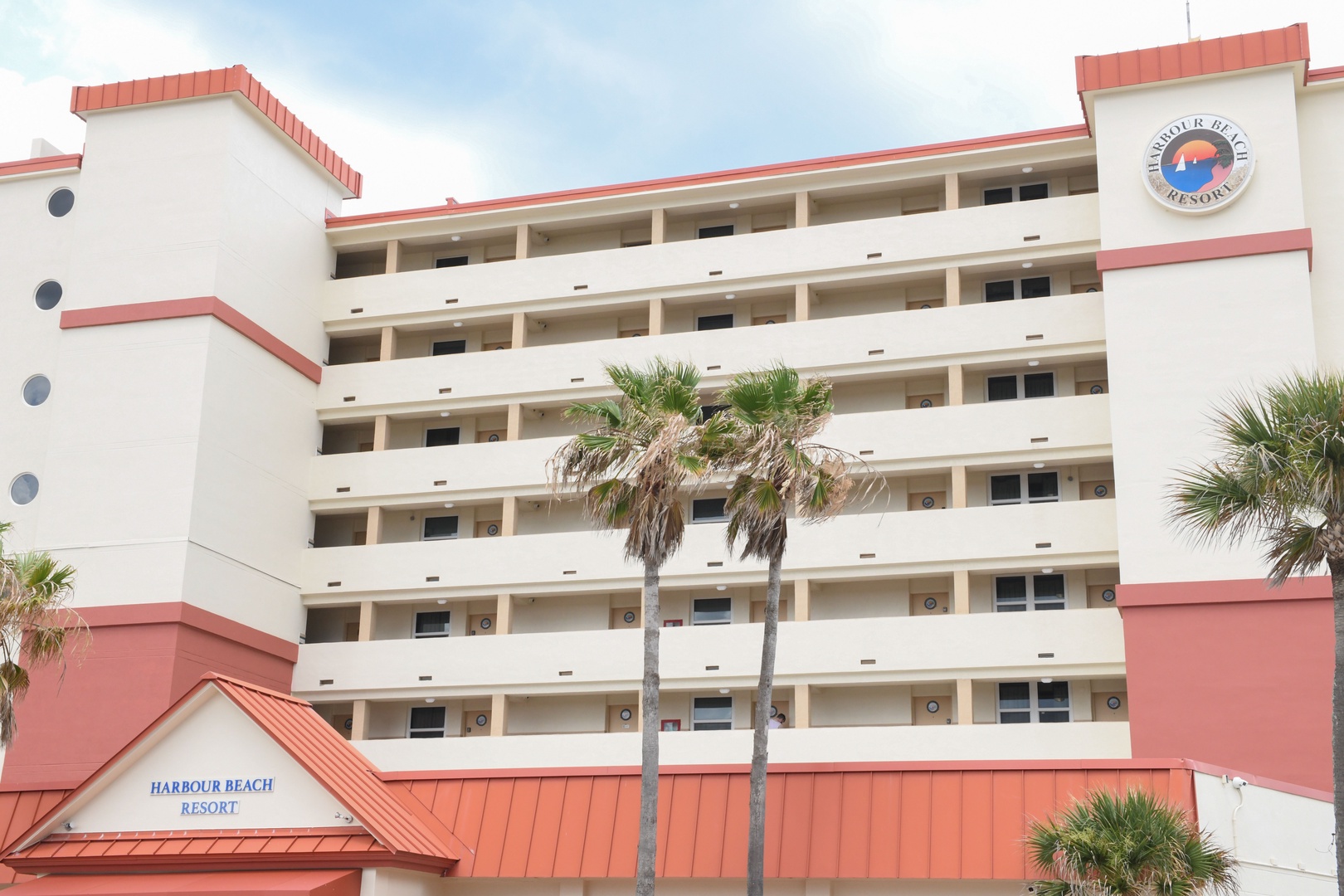 Harbour Beach Resort stands near the coast, featuring six stories of accommodations with coral trim and palm trees framing the property entrance.