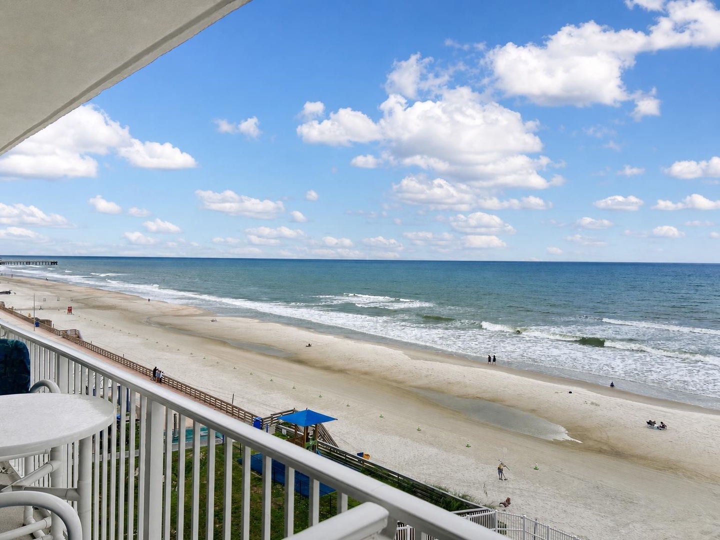 Sweeping oceanfront panorama stretching endlessly along pristine coastline, framed by gentle waves rolling onto soft sand under billowing clouds and brilliant blue sky.