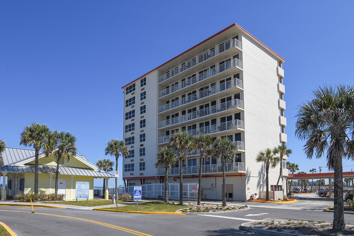 A modern beachfront property featuring multiple floors with private balconies, surrounded by tropical palm trees under clear blue skies.