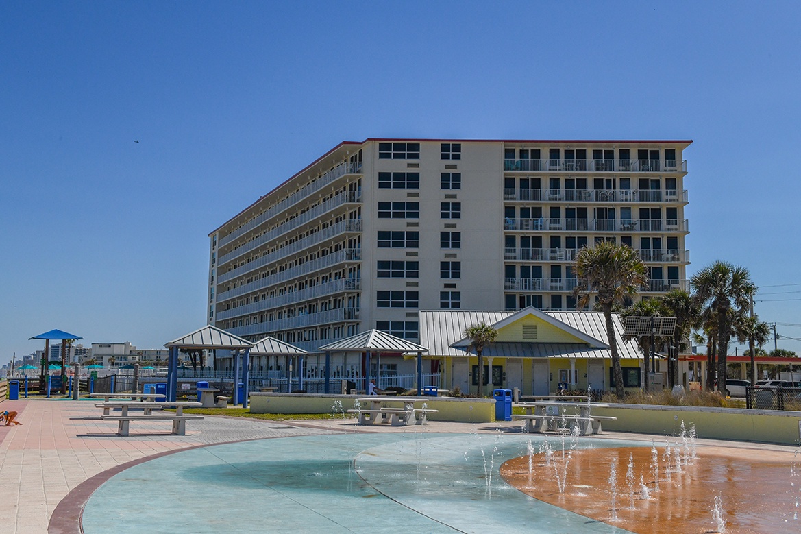 Beachfront property building with nearby splash pad and recreational facilities under clear blue skies.