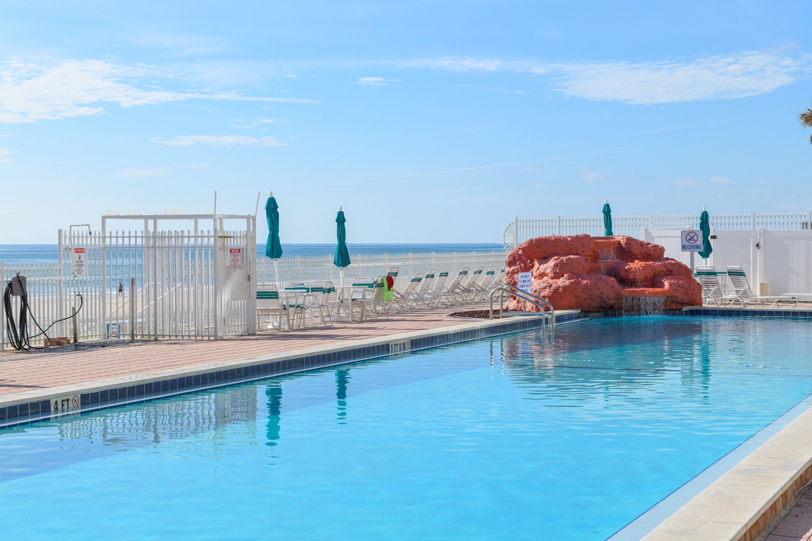 The pool area overlooks the ocean, featuring a unique rock formation waterfall, lounge chairs, and umbrellas for relaxation by the water.