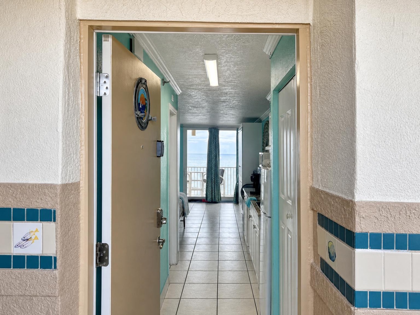 Step through the entryway and glimpse the open layout ahead—tile floors lead you straight to glass doors and a balcony framing the coastline. Calming teal accents set the mood for your coastal escape.