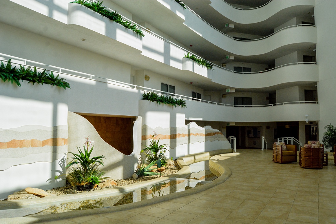 Step into this stunning atrium lobby where natural light cascades through multiple floors, creating a serene welcome with tropical plants and modern design.