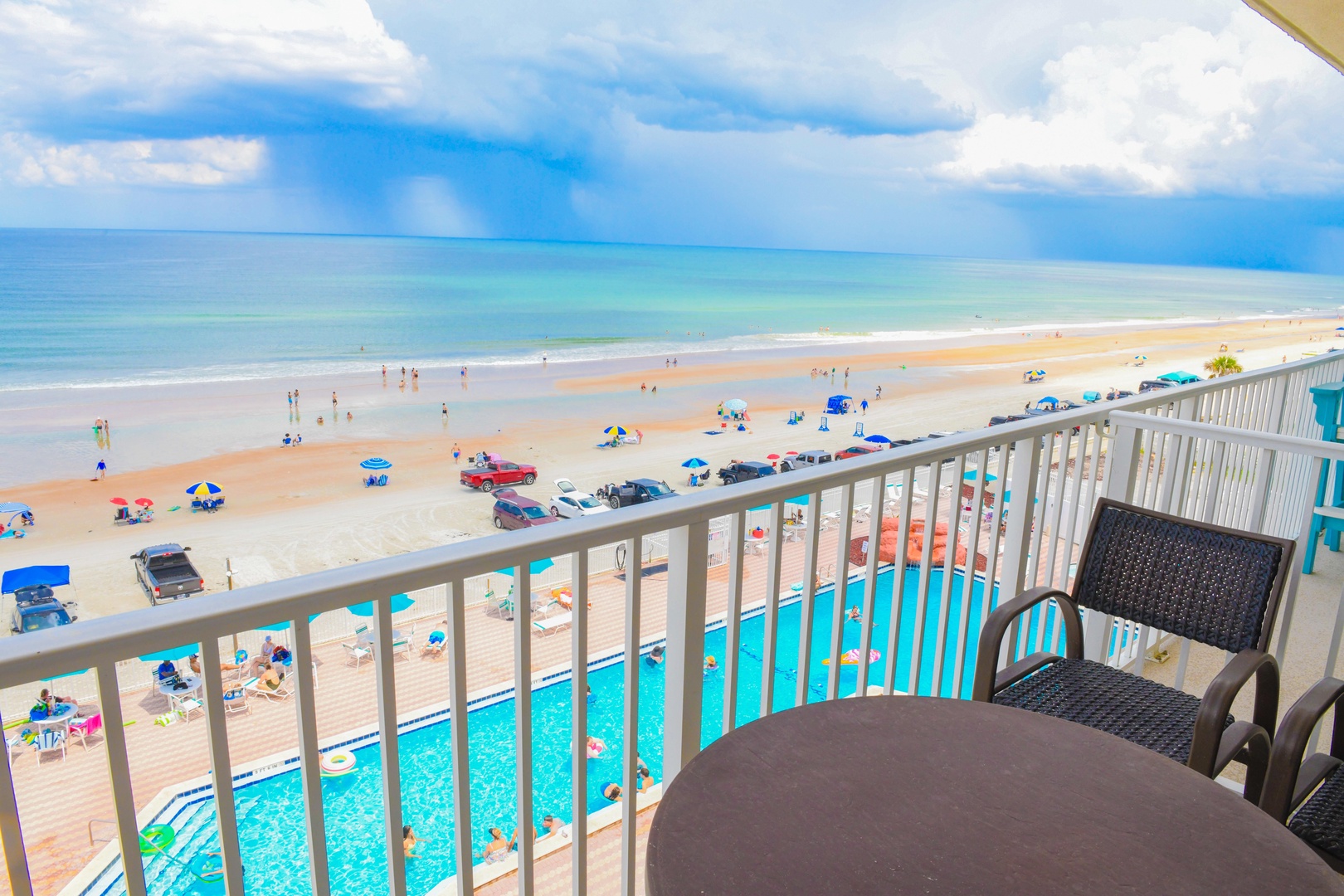 Expansive beachfront vista with direct ocean access, featuring a rectangular pool positioned between the building and sandy shore, with wide sandy beach extending to the water's edge under partly cloudy skies.